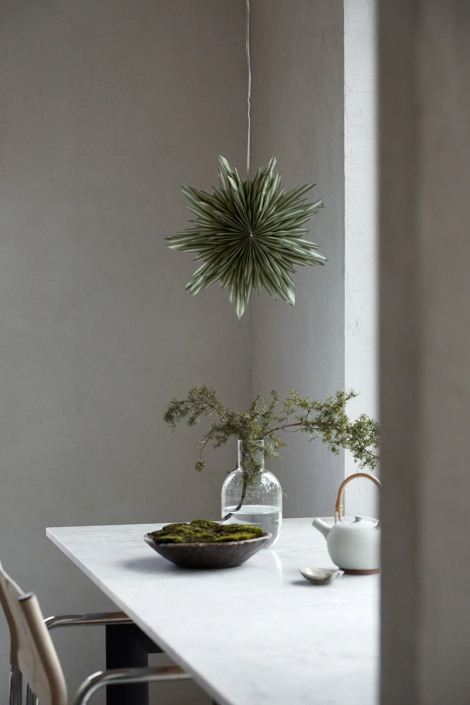 The paper star lantern Helena in green hangs in the window above a green plant in a pot. 