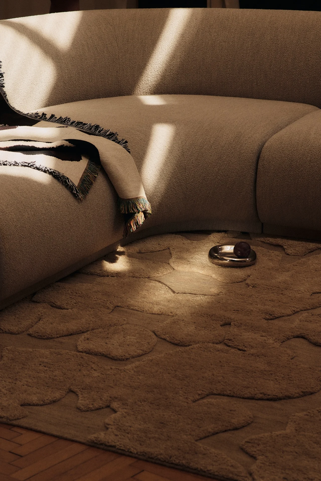 Curved beige sofa with a patterned throw, an organic-shaped beige rug, and a silver dish holding dark fruit in a sunlit room.