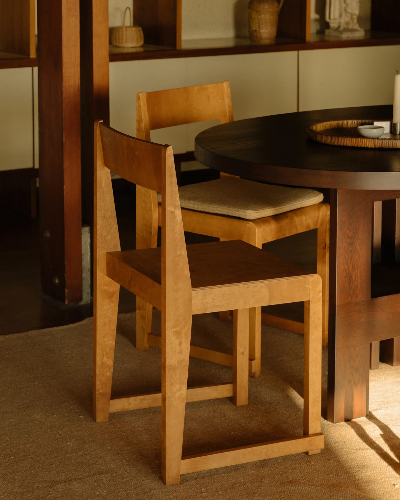 Two light wood chairs at a dark round dining table, with wooden shelves in the background.
