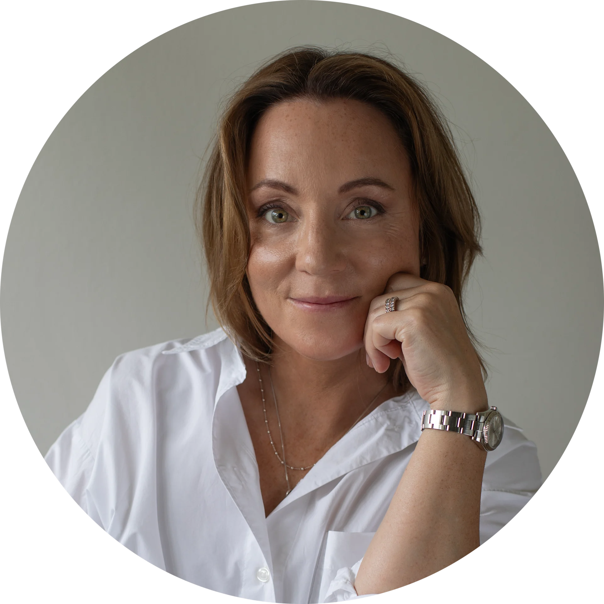 Woman with brown hair and green eyes in a white shirt, seated and smiling, resting her chin on her hand.