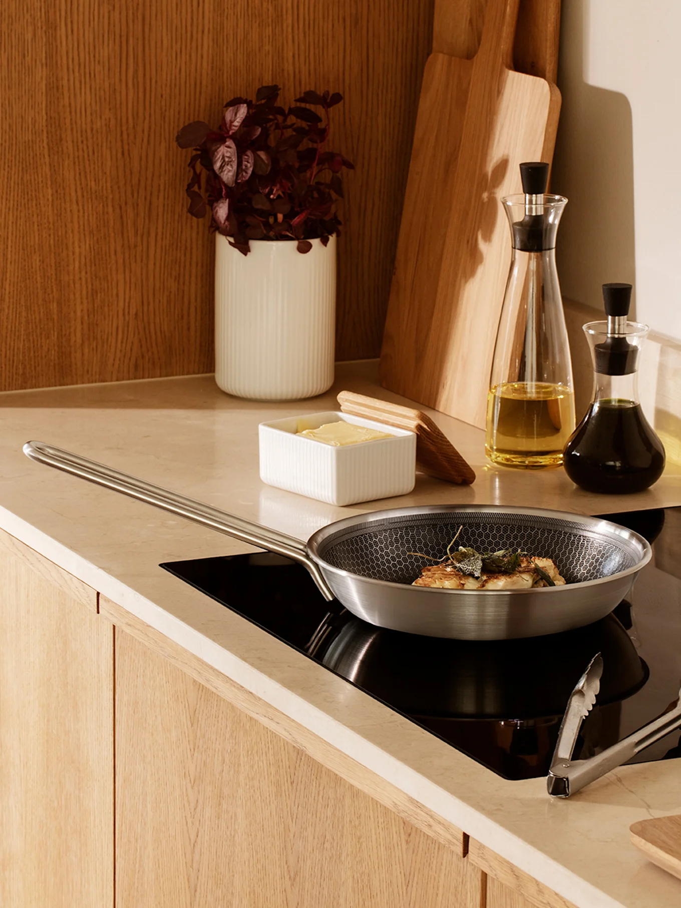A stainless steel frying pan with a honeycomb pattern cooking food with sage on a black induction hob in a modern kitchen.