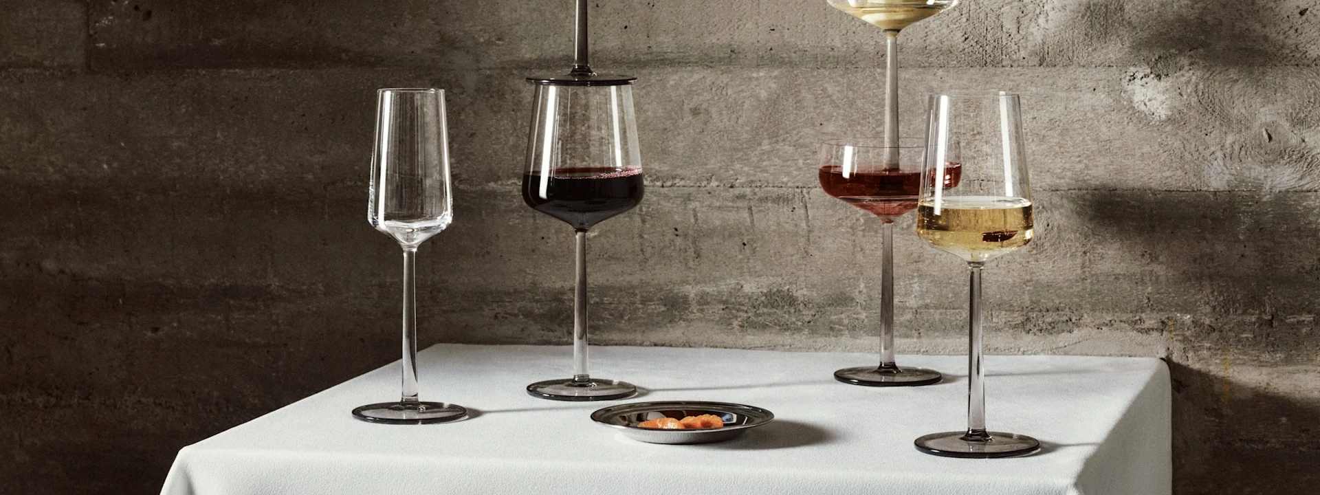Five elegant long-stemmed glasses with wine and champagne, one with a pear, on a white tablecloth in front of a concrete wall.