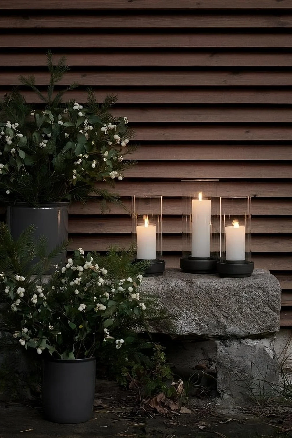 Three Hurricane lanterns from Stelton stand outside on stones, surrounded by plants.