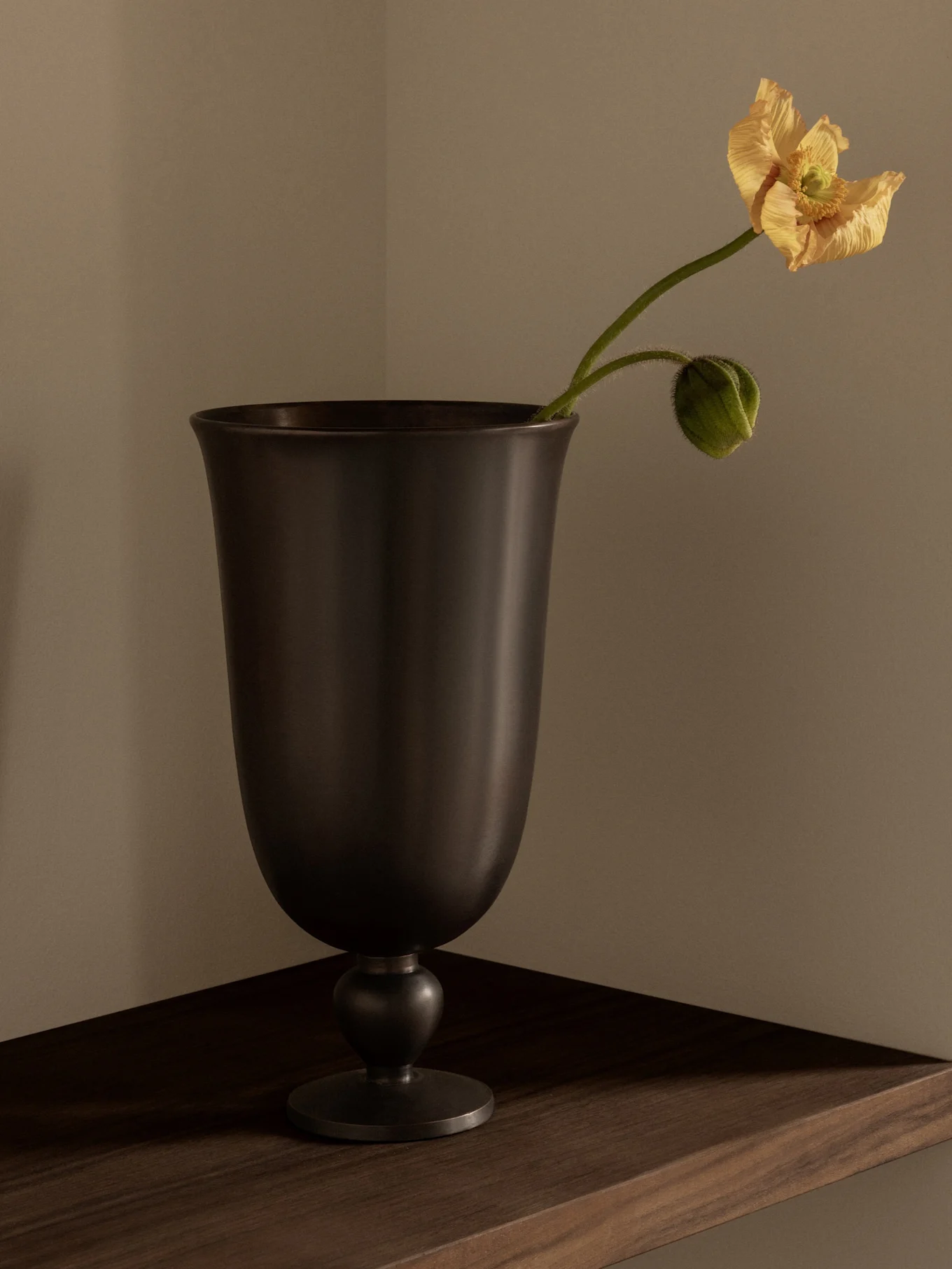 A dark, goblet-shaped vase holding a delicate yellow-orange poppy and a green bud on a wooden shelf.