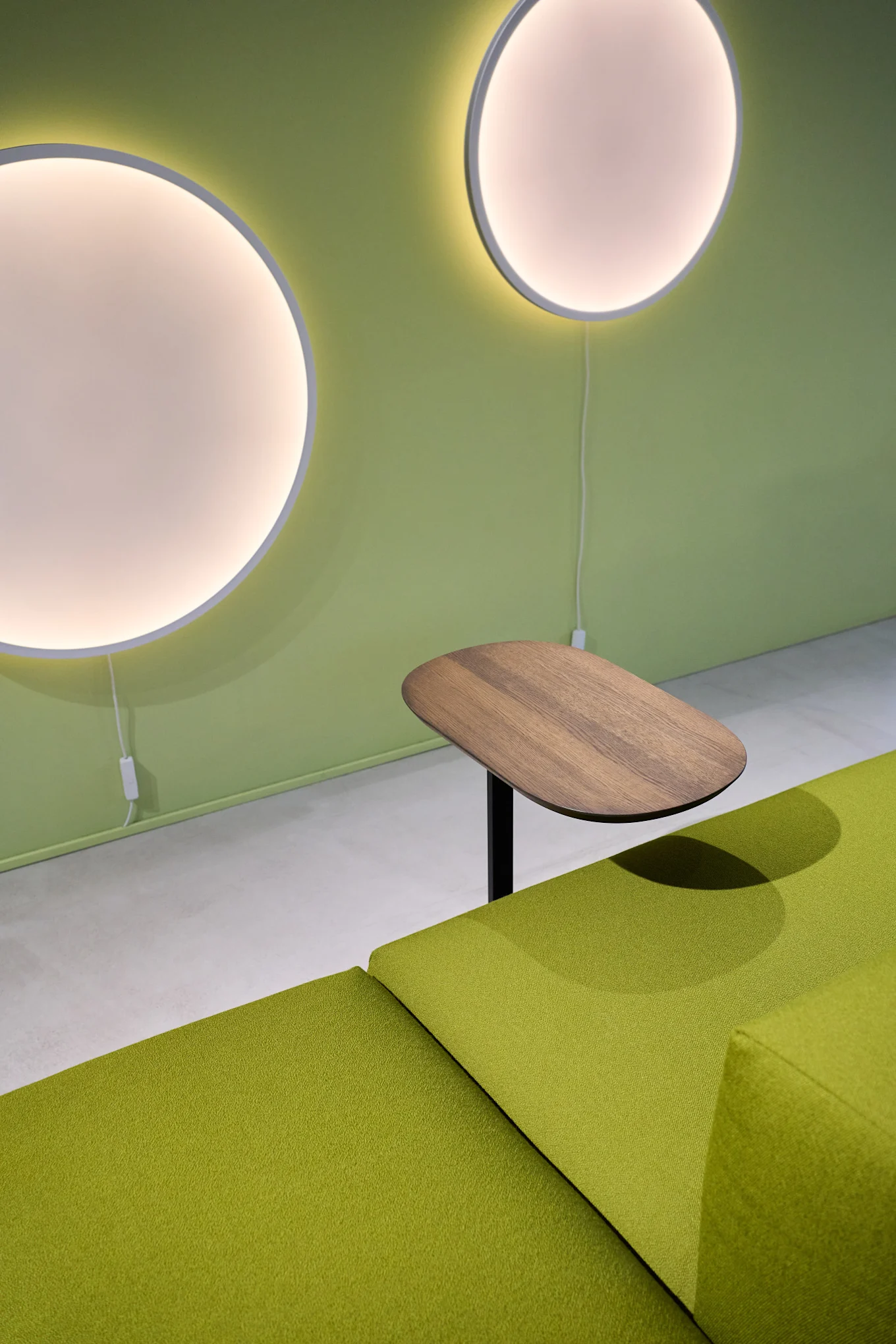 A room with a green wall on which two round Calm wall lights hang, in front of which is a green sofa with a Relate side table.