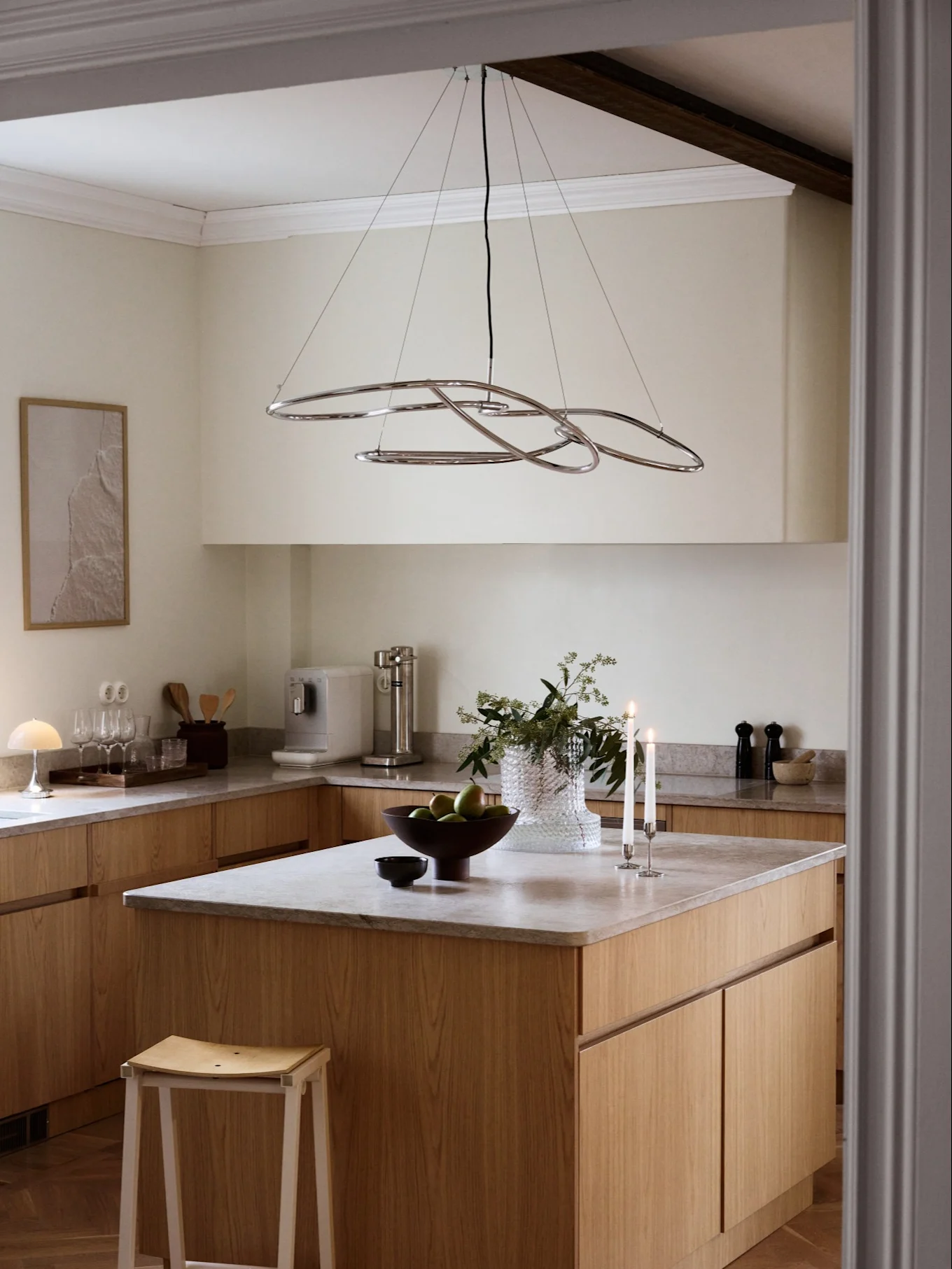 A modern kitchen with light wood cabinets, grey countertops, and a chrome abstract chandelier over an island with fruit and candles.