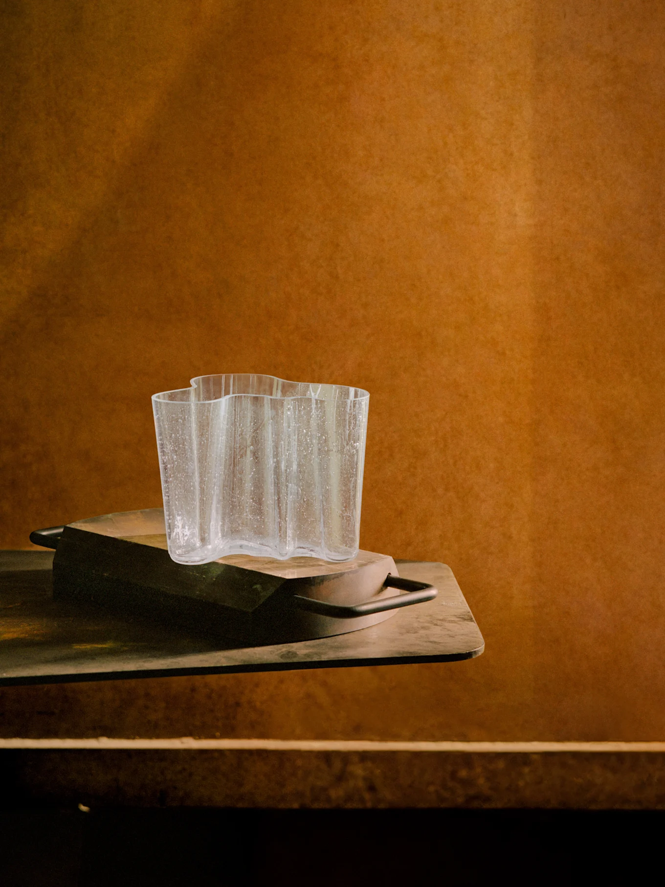 Clear, wavy glass vase with water droplets on a dark wooden tray, against a warm brown textured background.