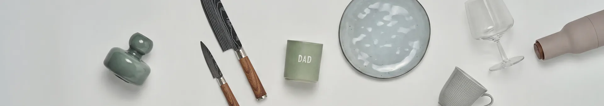 A flat lay of kitchen and gift items: a green vase, two knives, a 'DAD' candle, a grey ceramic plate, a wine glass, a beige bottle, and a grey mug.