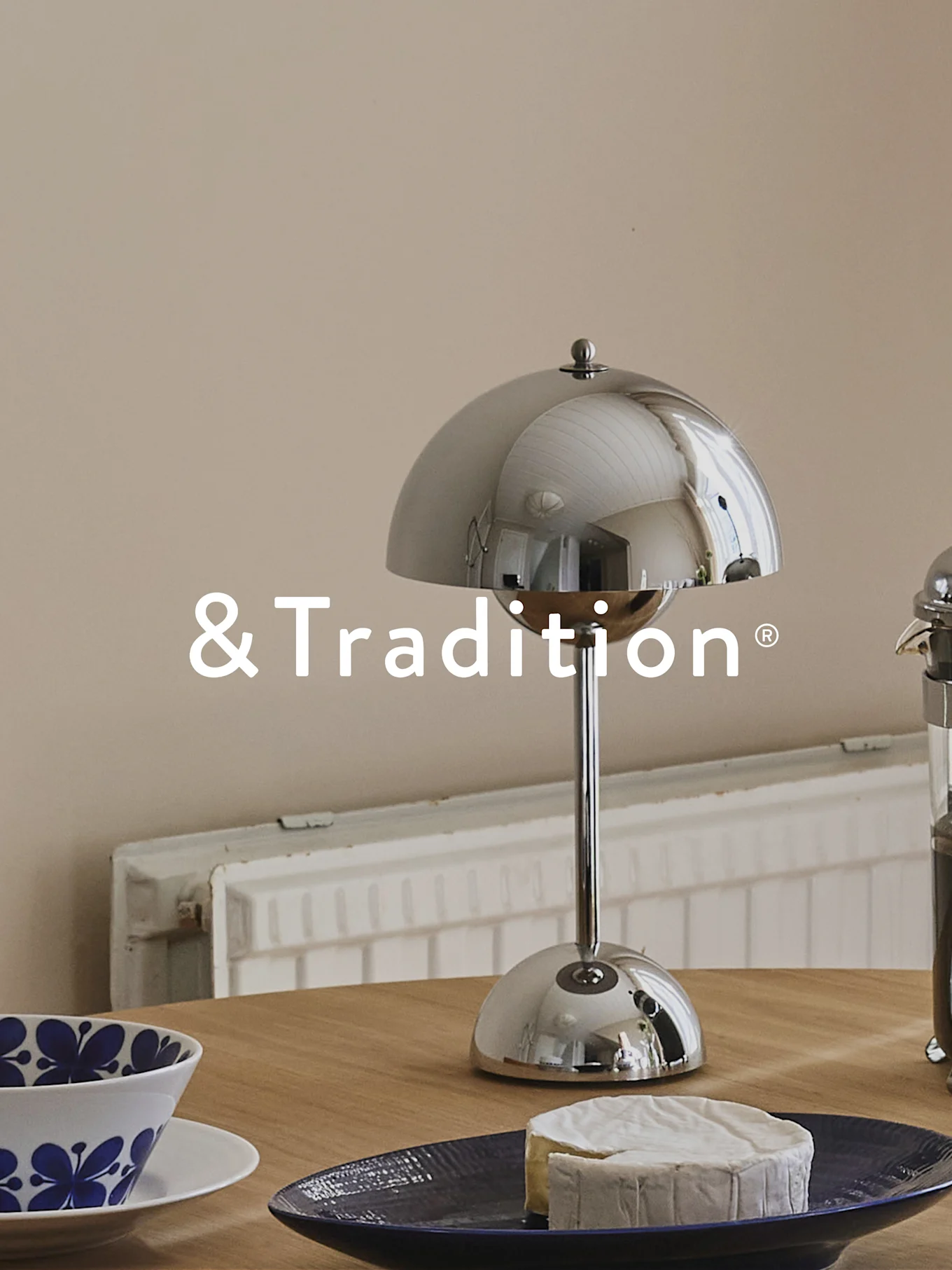 A silver dome-shaped table lamp, a round cheese on a dark blue plate, and a blue patterned bowl on a wooden table. &Tradition logo.