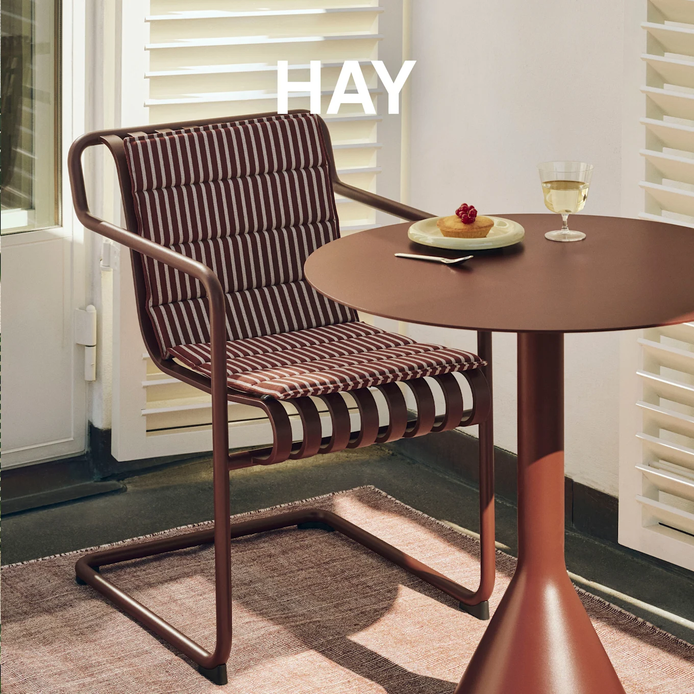 Reddish-brown outdoor chair with striped cushions and matching table with cake and wine on a rug. HAY logo visible.