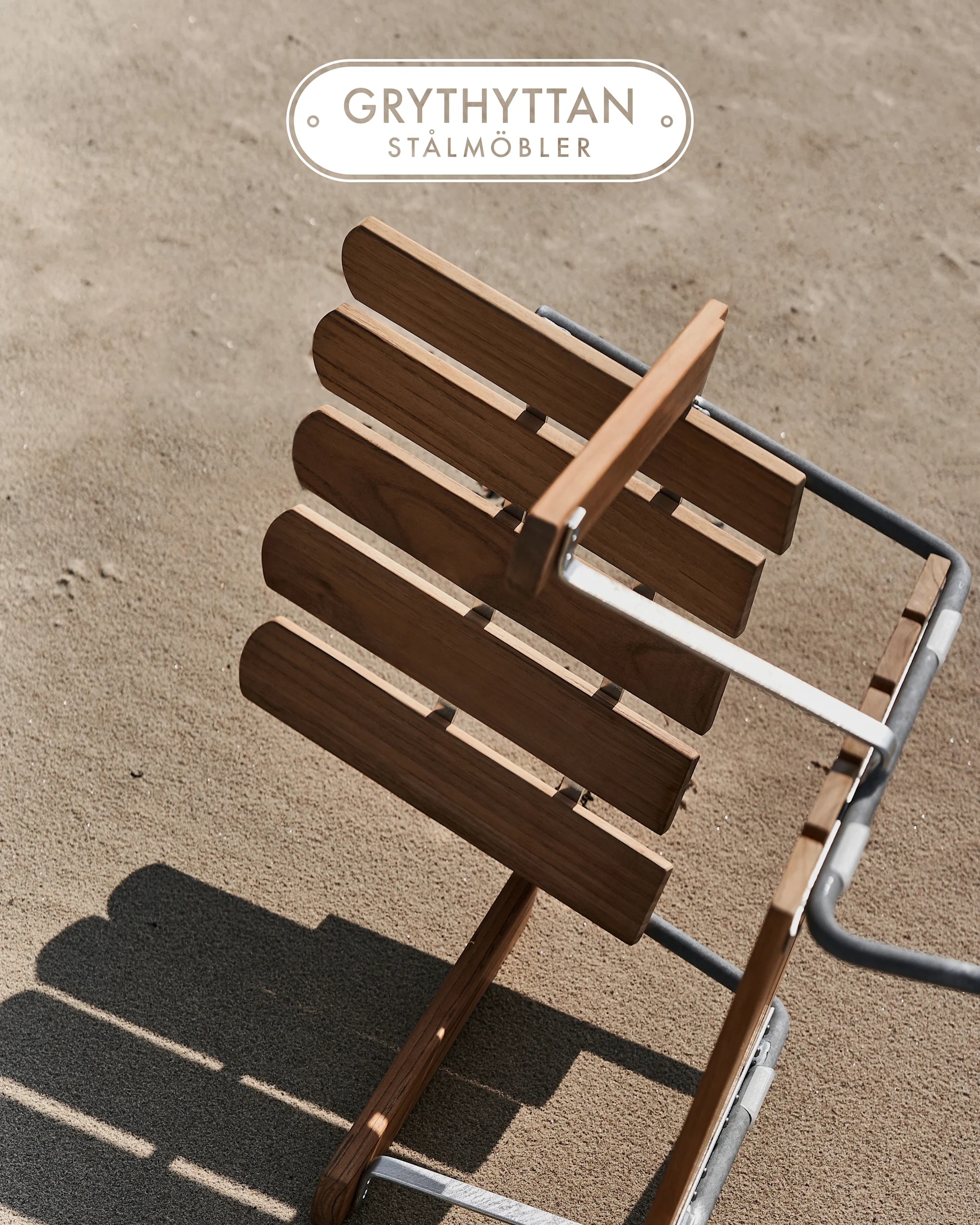 Wooden slatted chair with metal frame on sand, seen from above. Grythyttan Stålmöbler logo.