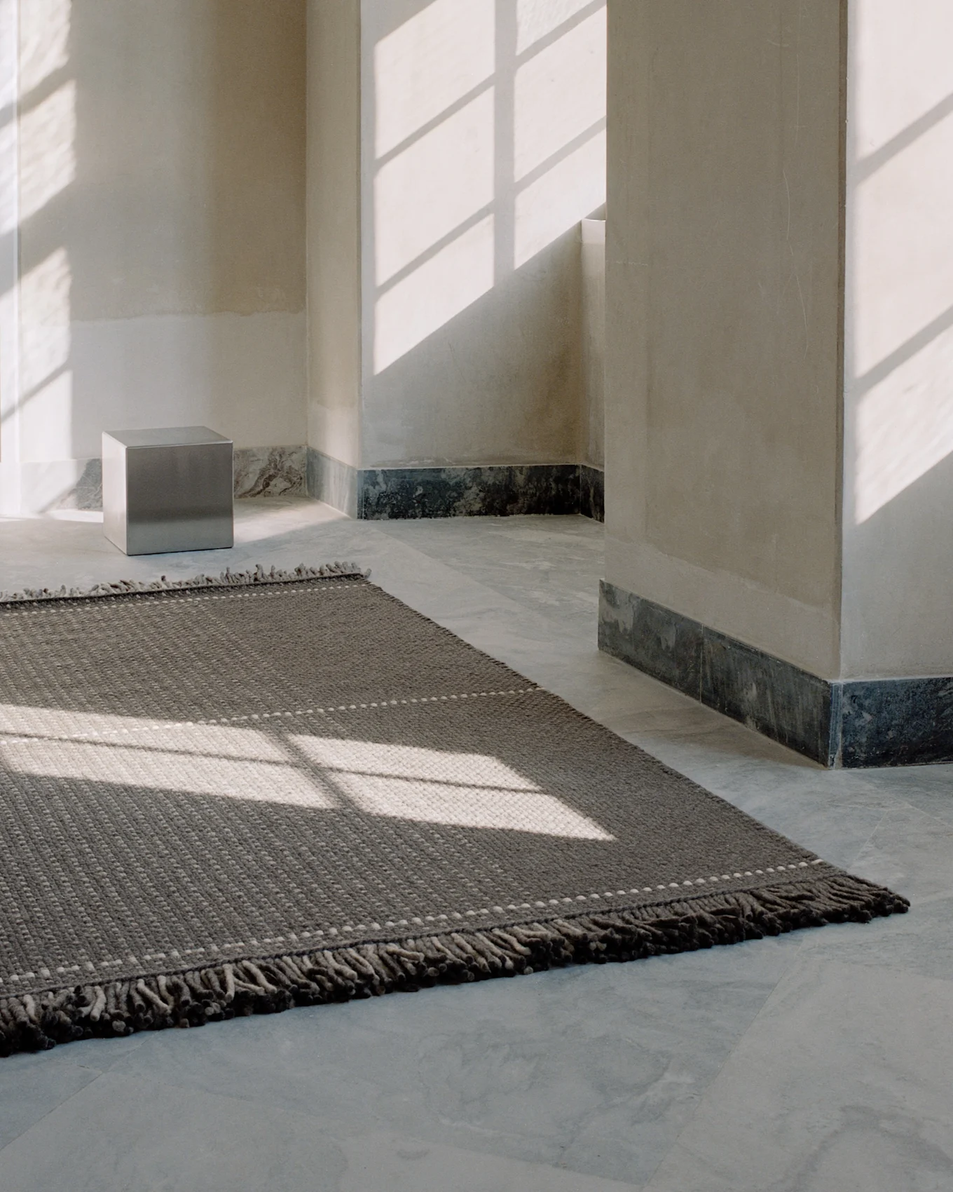 A dark gray textured rug with light pattern dots on a light tiled floor, illuminated by window shadows in a minimalist room.