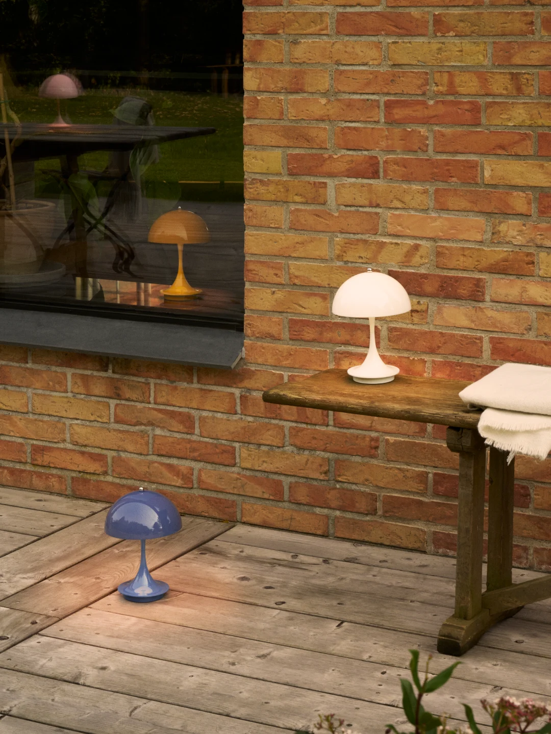 White mushroom lamp on table, blue on deck. Pink and yellow mushroom lamp reflections in window by brick wall.