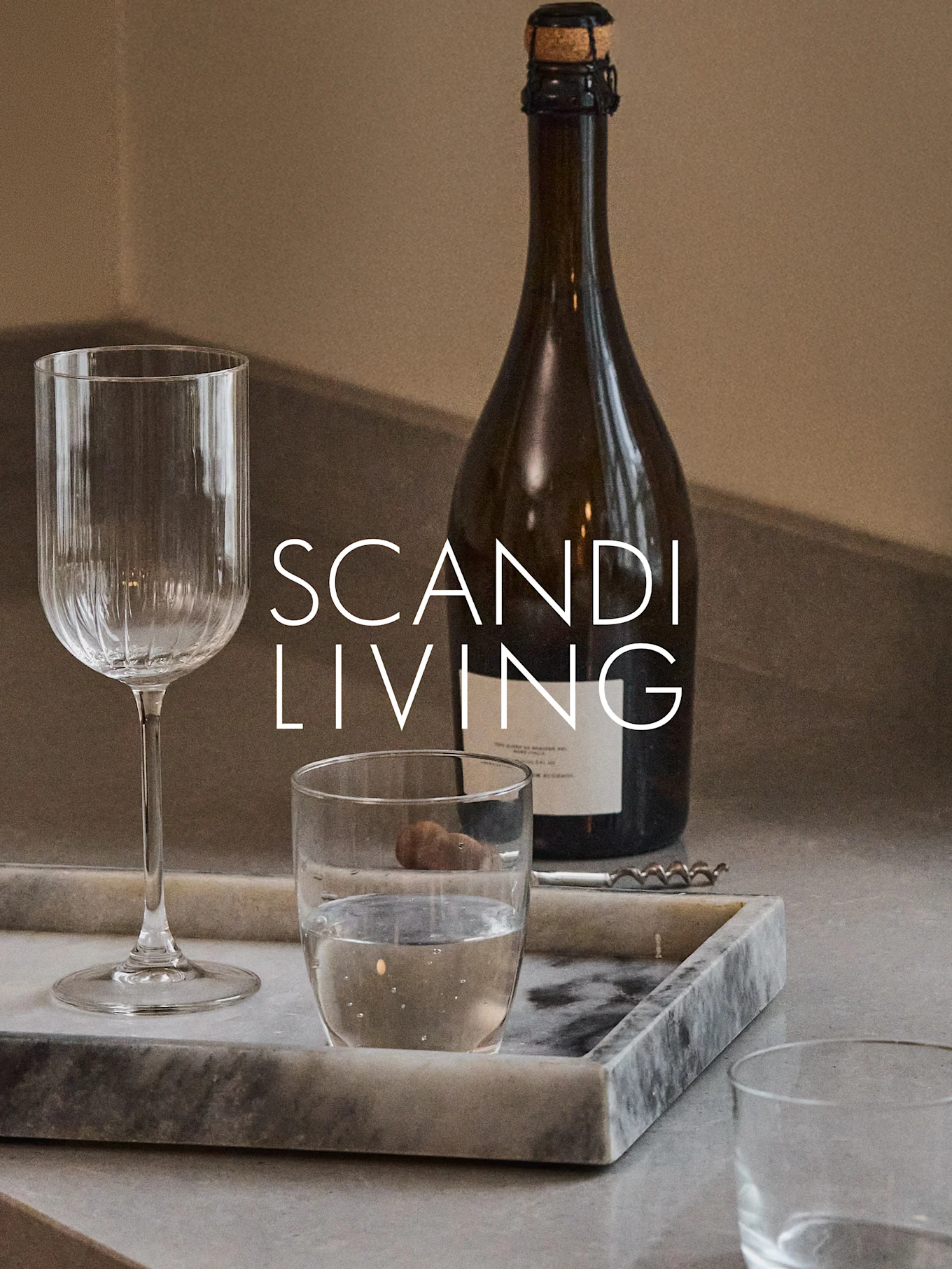 A dark bottle, a ribbed wine glass, a water glass, and a corkscrew on a marble tray with 'SCANDI LIVING' text.