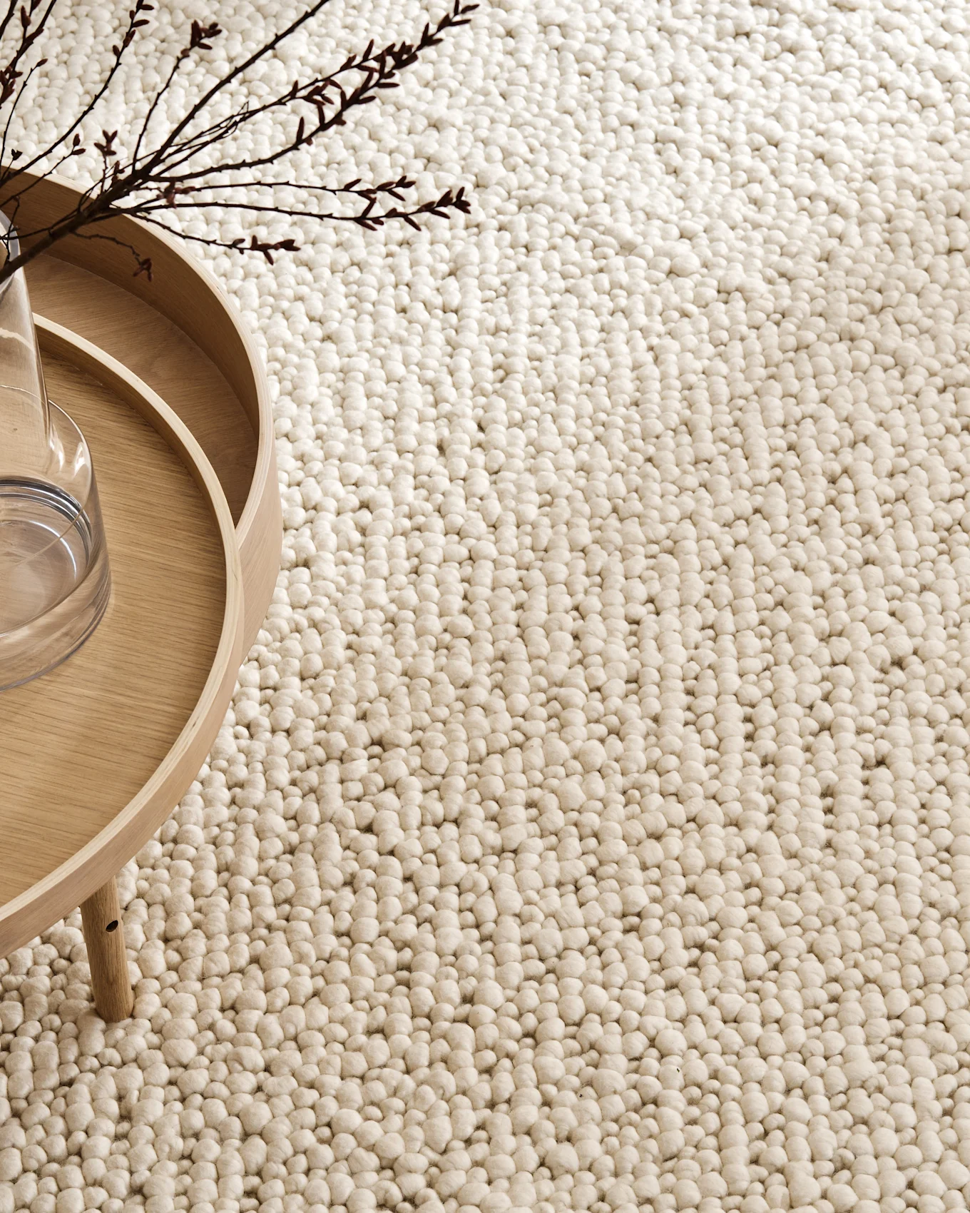 Textured light beige rug with a bumpy, bobble-like surface; a wooden table and bare branches are partially visible.