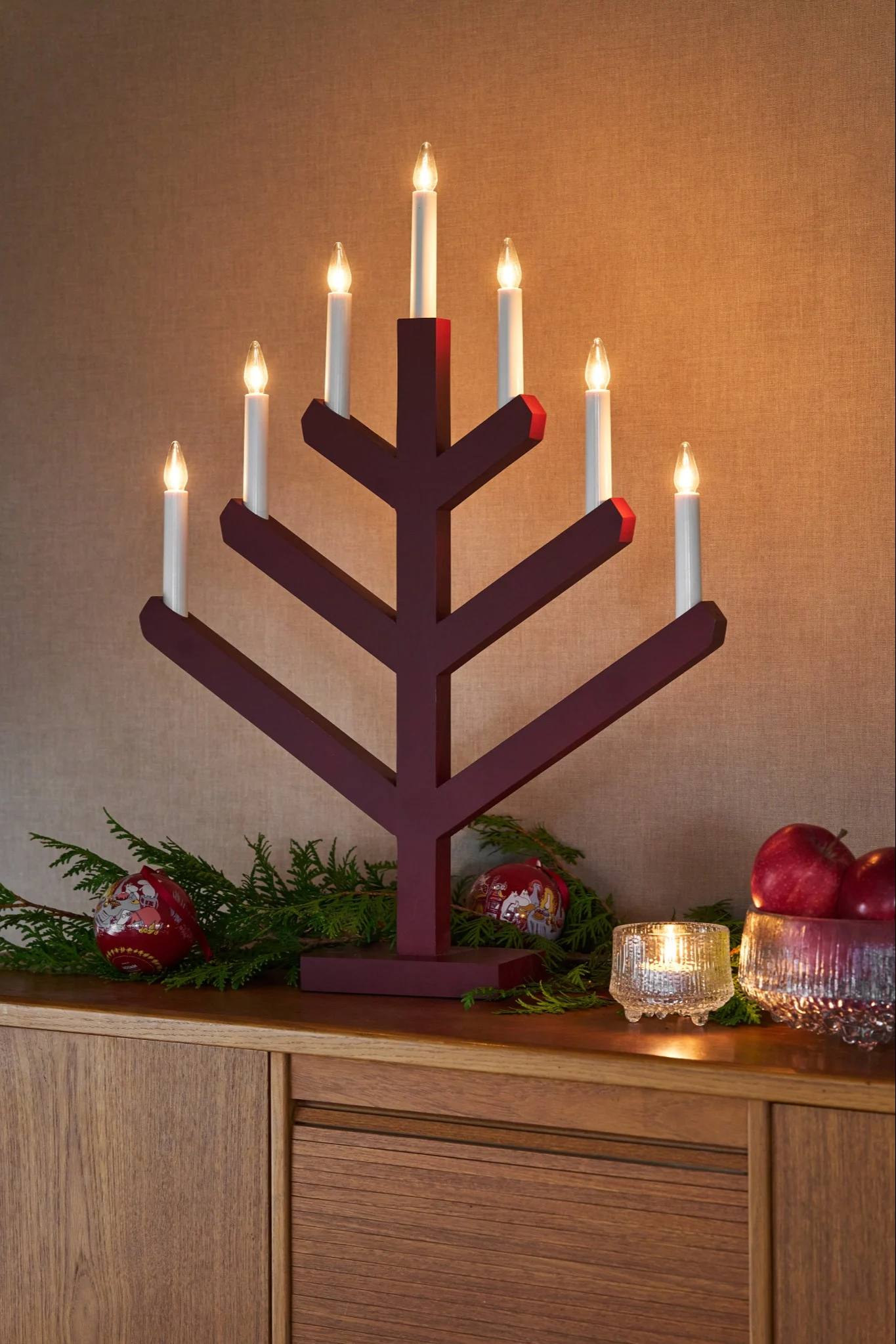 A red Advent candle holder shines beautifully on a sideboard alongside spruce branches and glittering Christmas baubles.