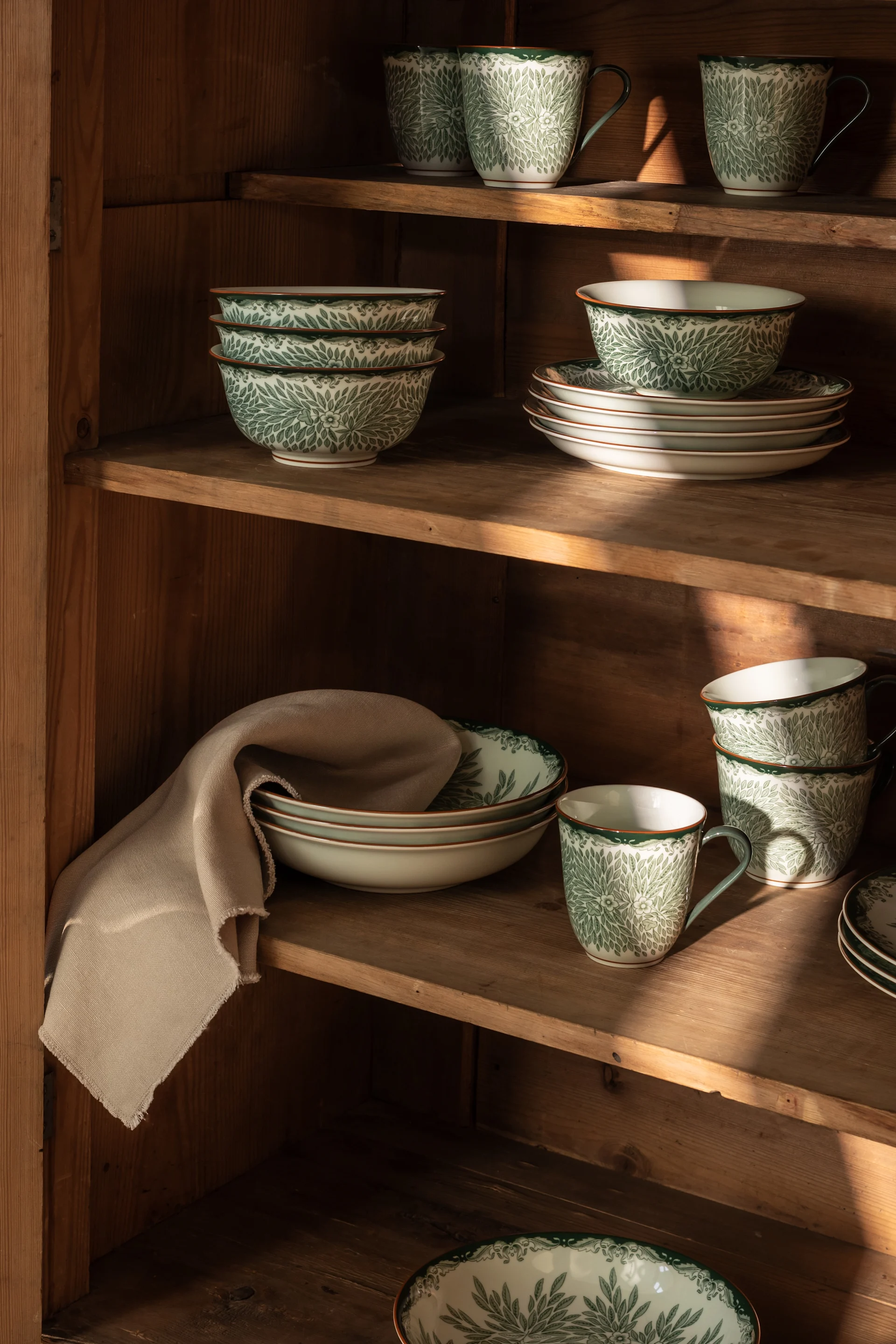 White and green patterned dinnerware, including bowls, plates, and mugs, displayed on rustic wooden shelves with sunlight.
