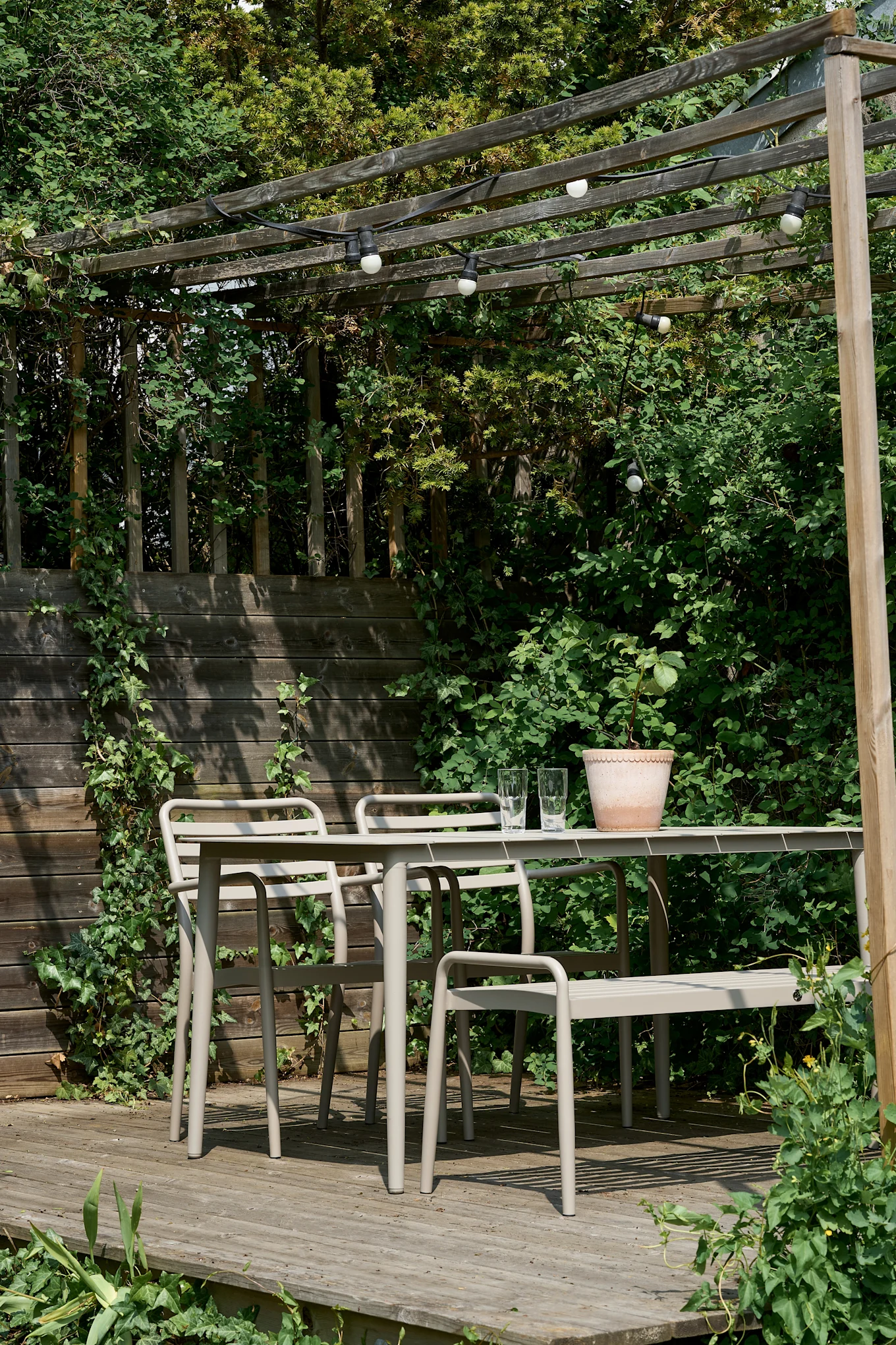An outdoor dining set with a beige metal table and chairs on a wooden deck under a pergola with string lights.