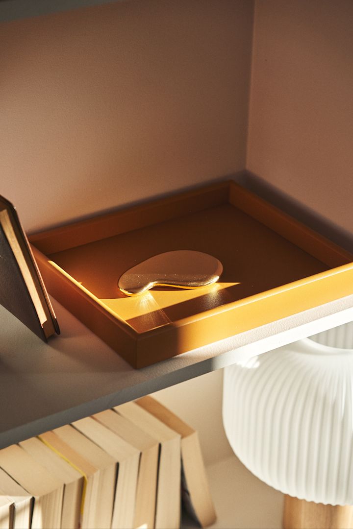 A dark mustard coloured tray stands on a wooden shelf in the sunshine. The tray is an ideal way to organise the living room. 