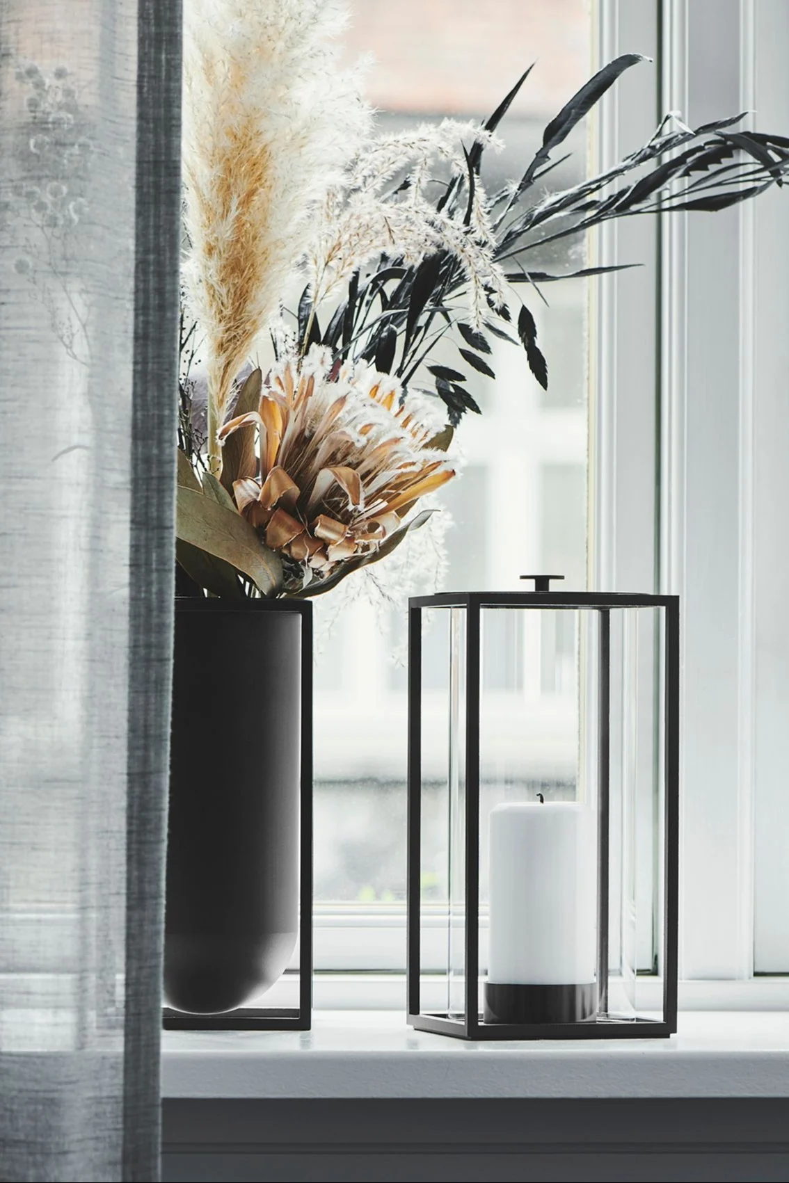 The Light'In lantern from Audo Copenhagen is perfect for decorating your windowsill, especially in the cooler months of the year. Here you can see it with a block candle next to a vase of dried flowers on the windowsill.