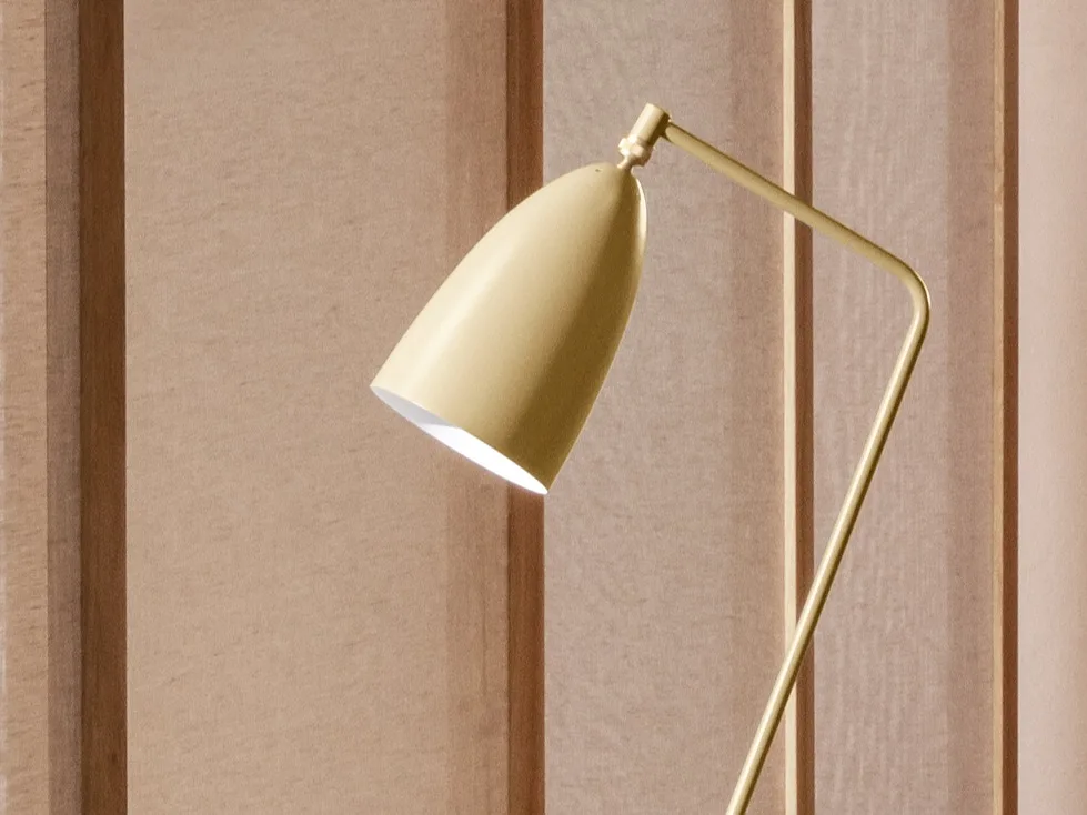 A close-up of a yellowish-gold modern lamp with a conical head angled down, emitting light against a light brown paneled wall.