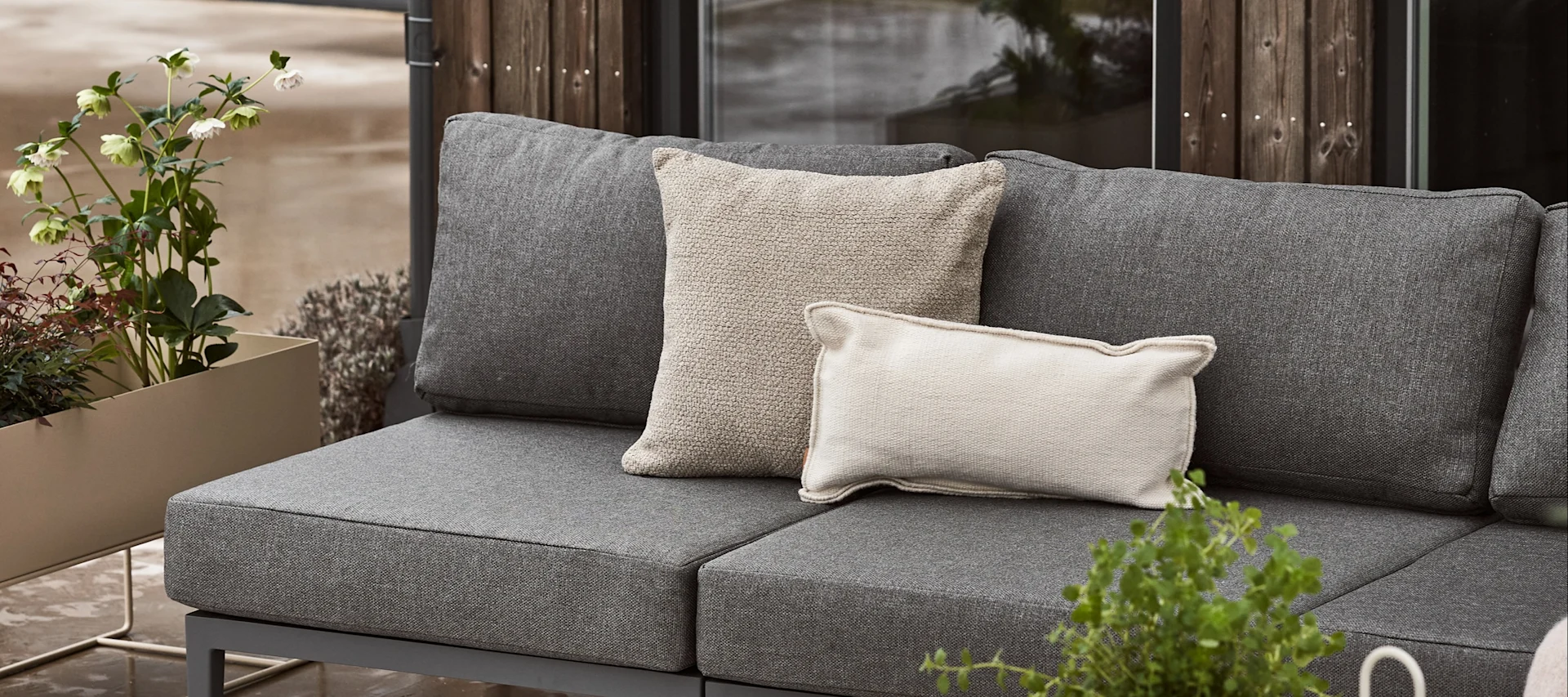 A grey outdoor sofa with two light-colored throw pillows, next to a planter with green plants and white flowers.