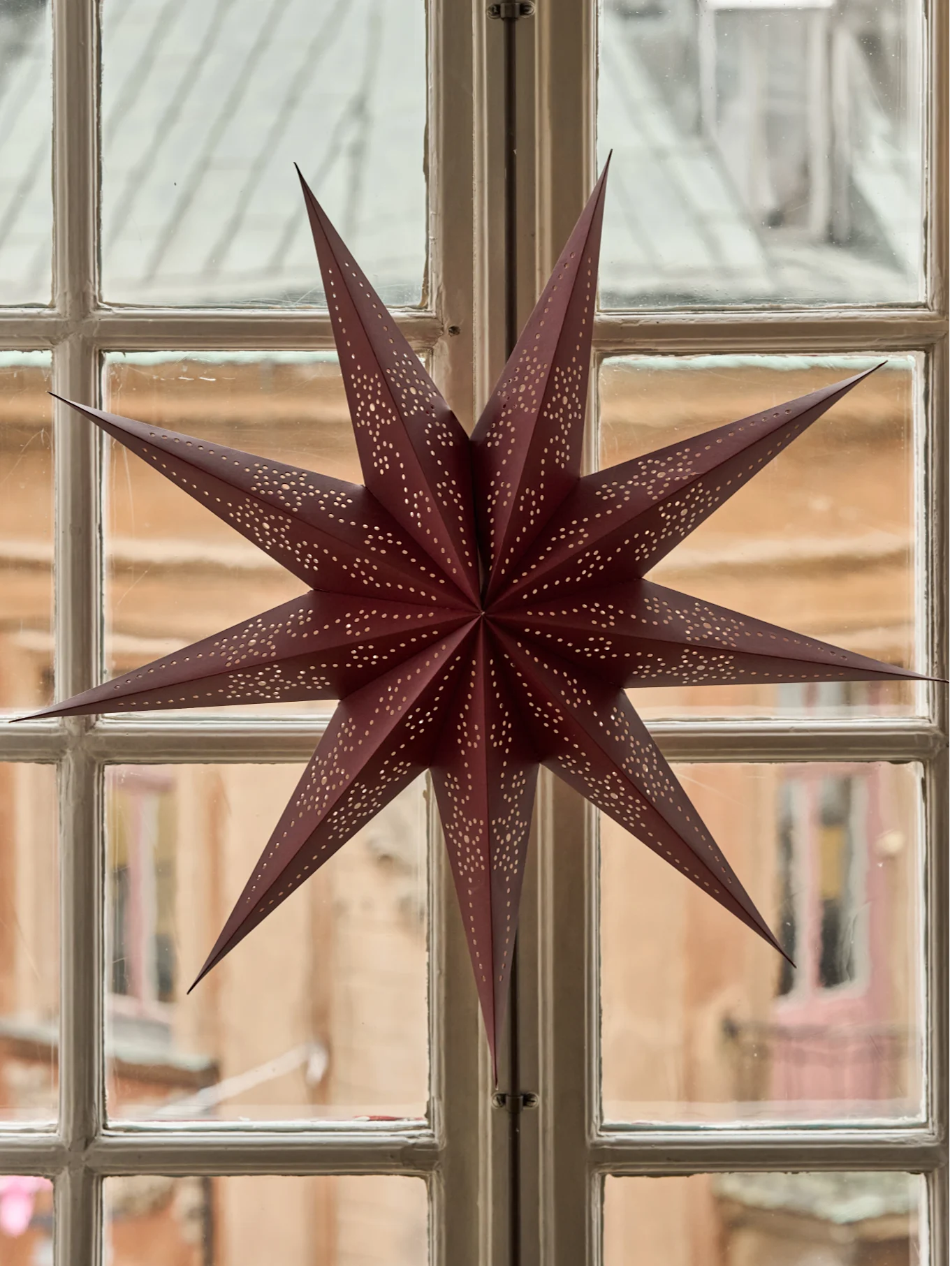 A maroon paper star with punched-out holes hangs in a multi-paned window, blurring the buildings outside.