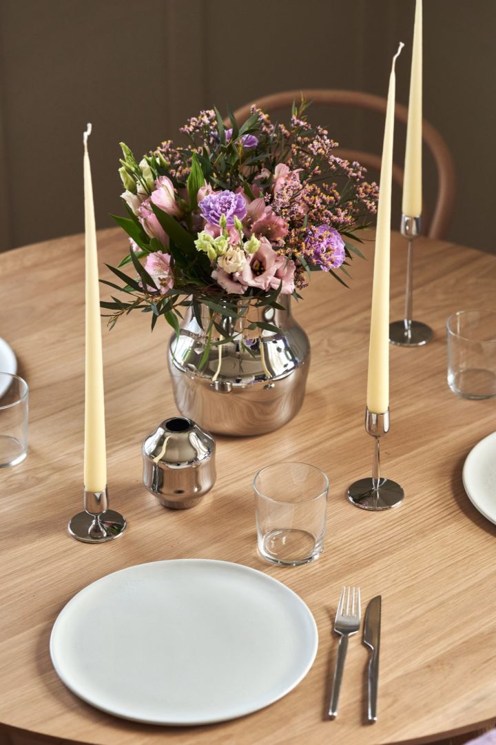 A mirrored stainless steel vase stands in the middle of a table with a bouquet of fresh purple flowers. 