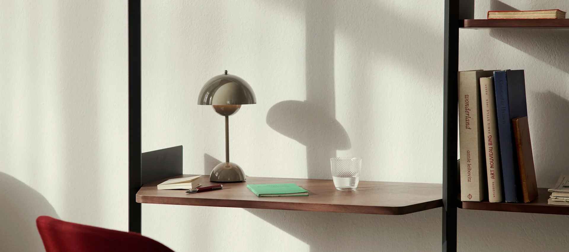 A modern mushroom-shaped desk lamp, a glass of water, and notebooks on a wooden desk shelf next to a bookcase.