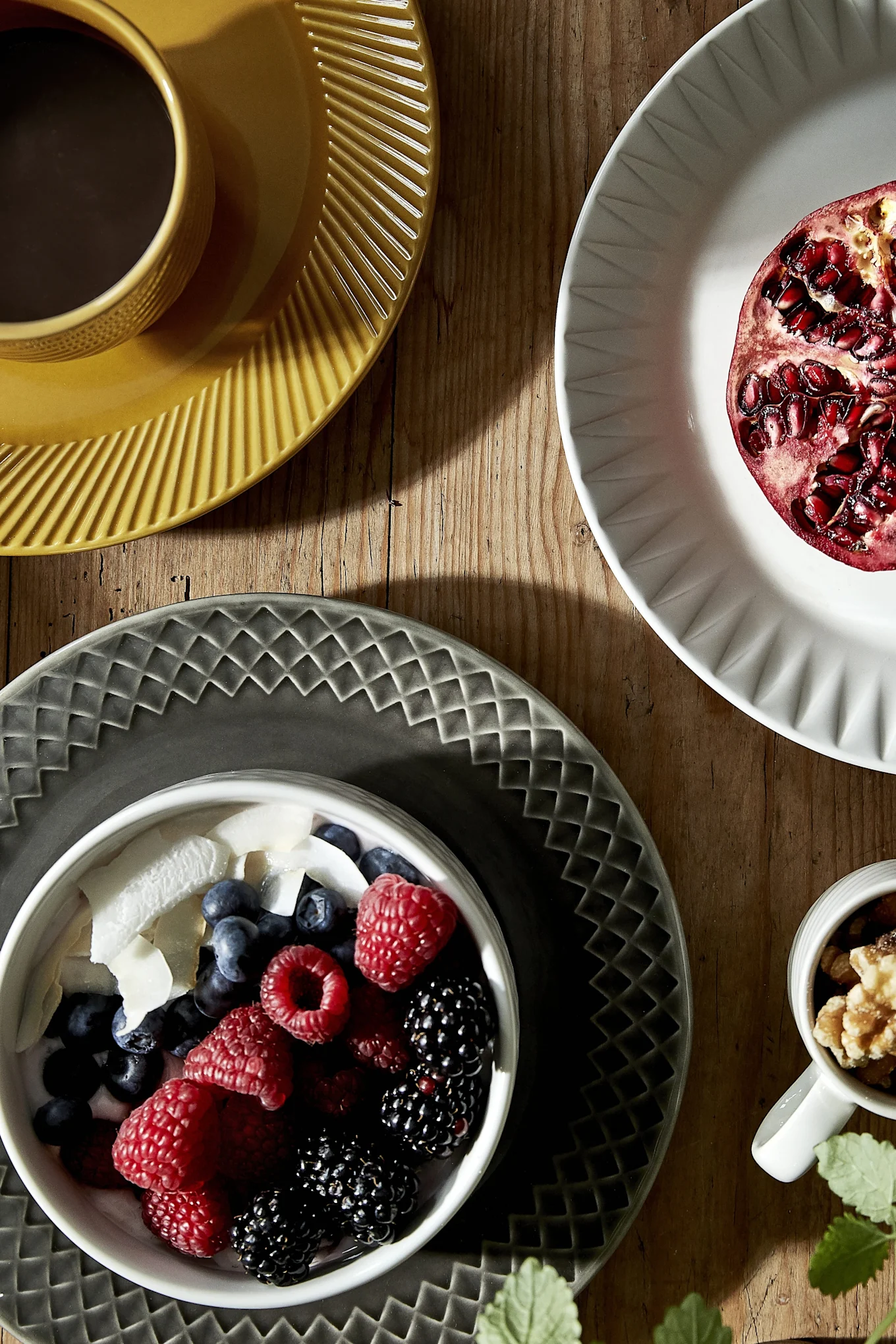 Overhead shot of breakfast on a wooden table with a bowl of berries and yogurt, walnuts, a pomegranate half on a white plate, and coffee in a yellow mug.