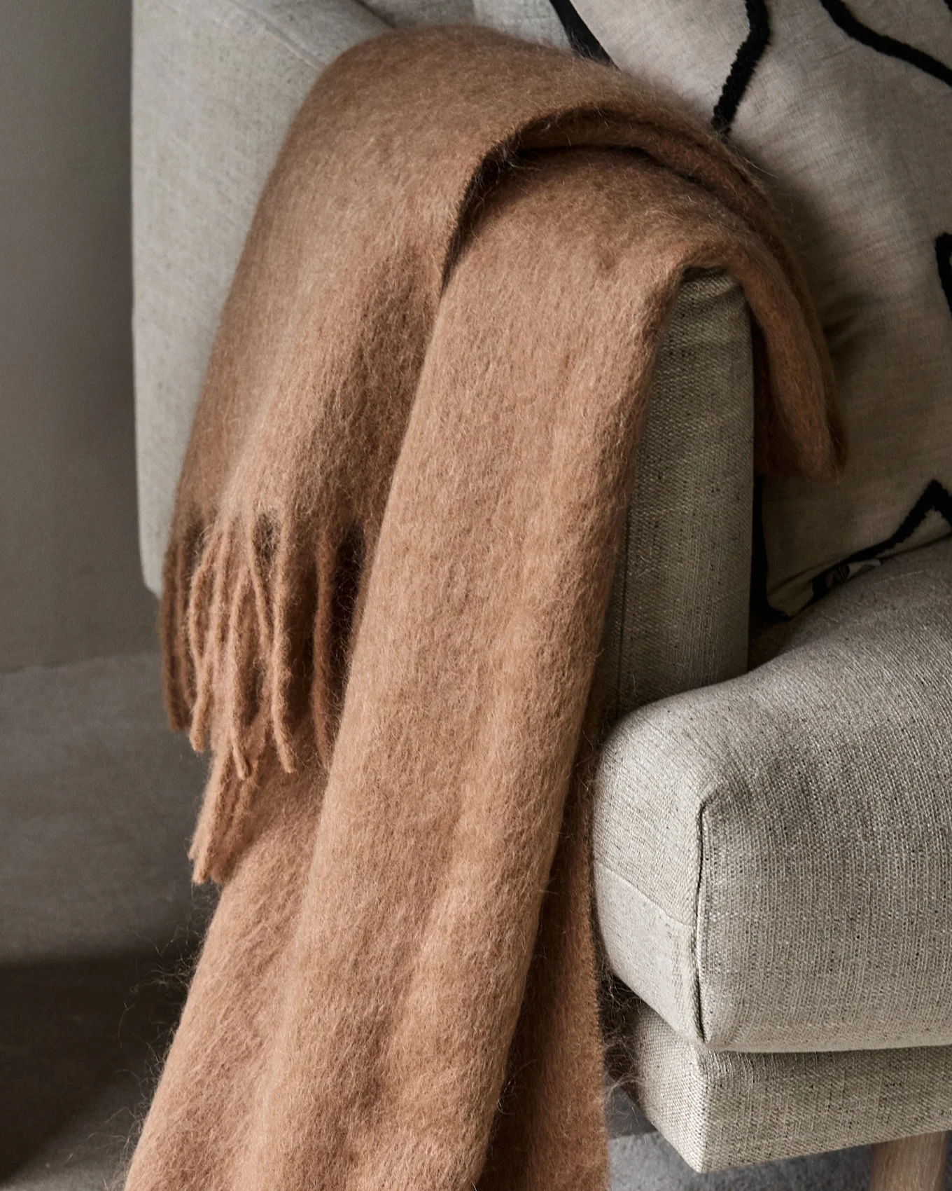 Cozy light brown fluffy throw blanket with fringes draped over a light grey armchair.