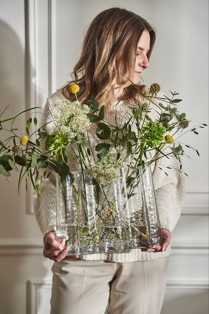 A woman in cream stands holding the Gry vase wide filled with wild spring flowers. 