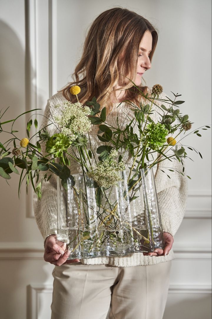 A woman in cream stands holding the Gry vase wide filled with wild spring flowers. 