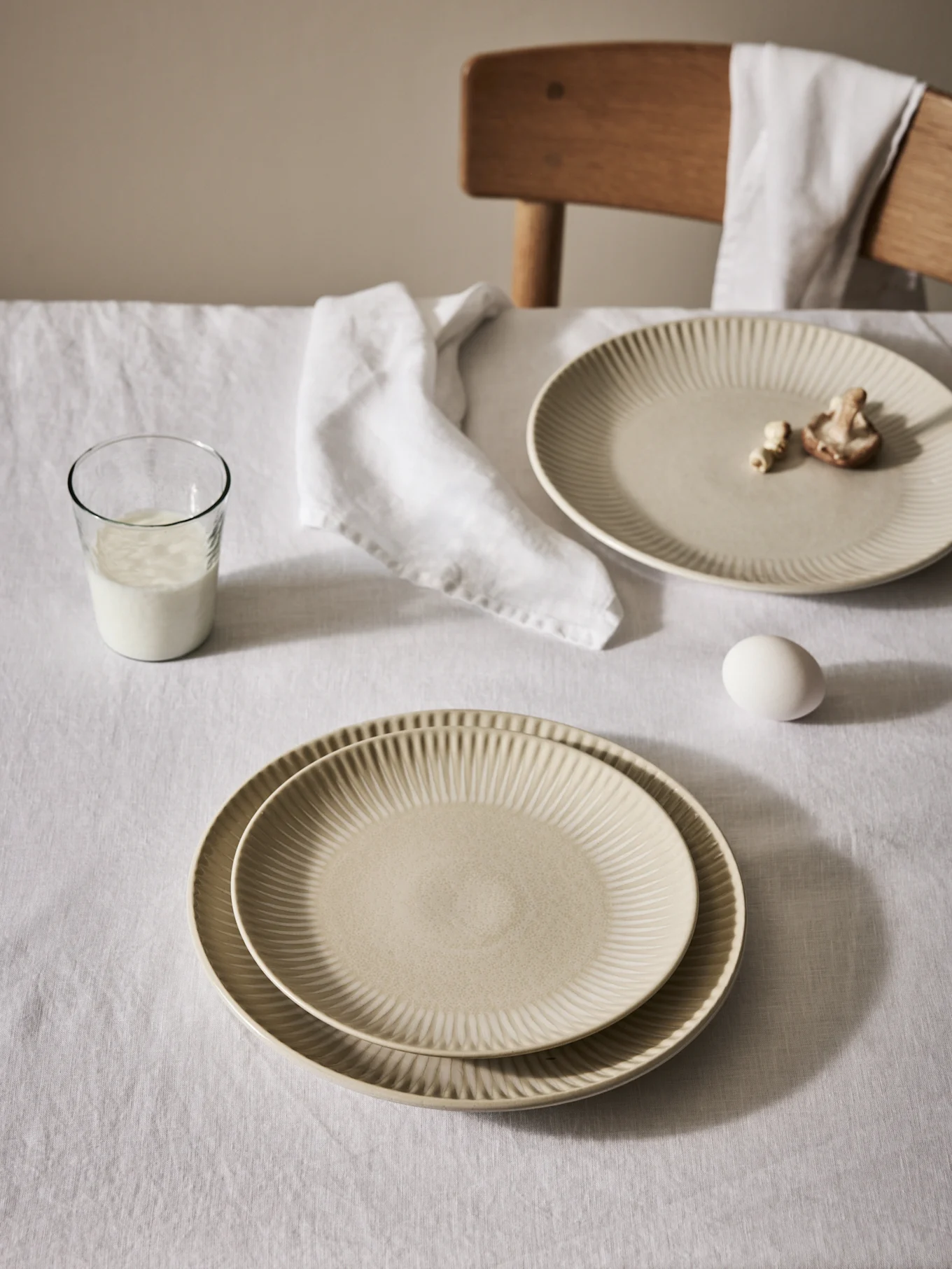 Table set with beige ribbed plates, a glass of milk, an egg, and a napkin on a white tablecloth.