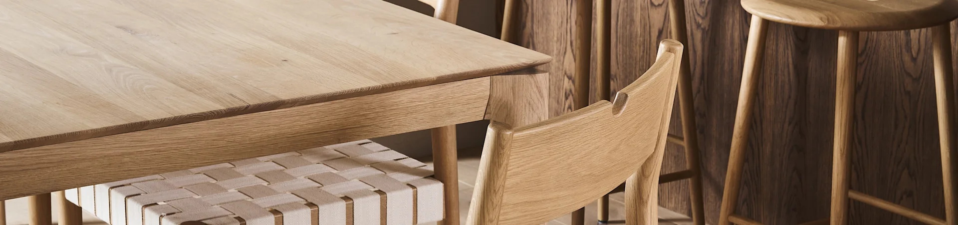 Close-up of a light wooden dining table with a chair featuring a woven seat and wooden stools in the background.