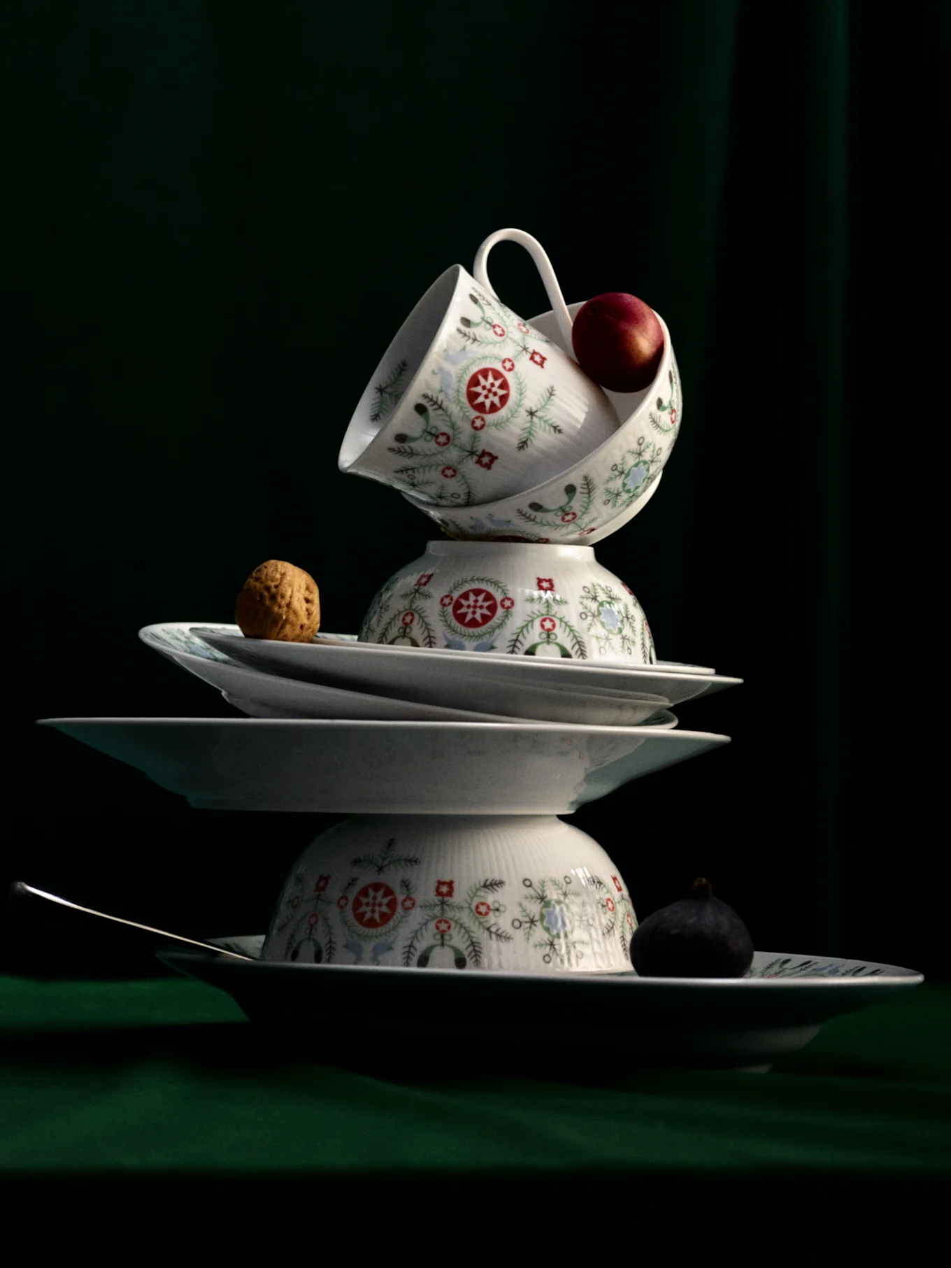 Still life of stacked white dinnerware with festive patterns, featuring an apple, walnut, fig, and spoon on a dark green background.