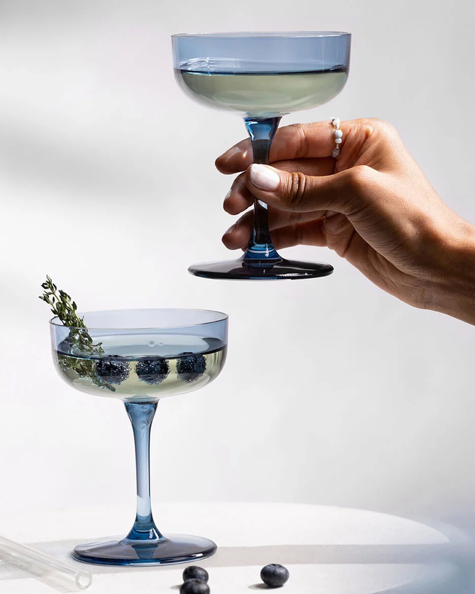 A hand holds a blue coupe glass with a drink, while another sits on a table with blueberries and thyme.