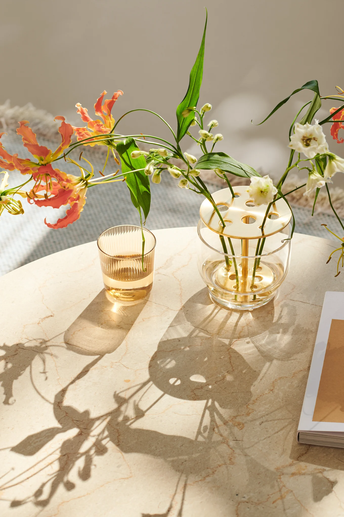 Floral arrangement with orange Gloriosa lilies and white flowers in a modern vase, beside a fluted glass, on a marble table with sunlight and shadows.