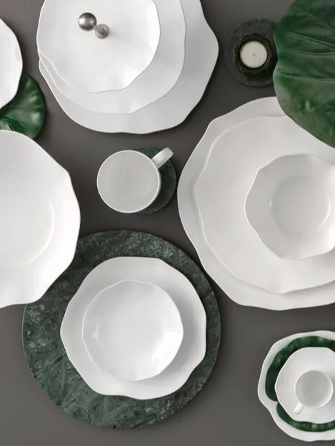 A collection of white ceramic dinnerware with wavy edges, arranged with green leaves and marble coasters on a dark surface.