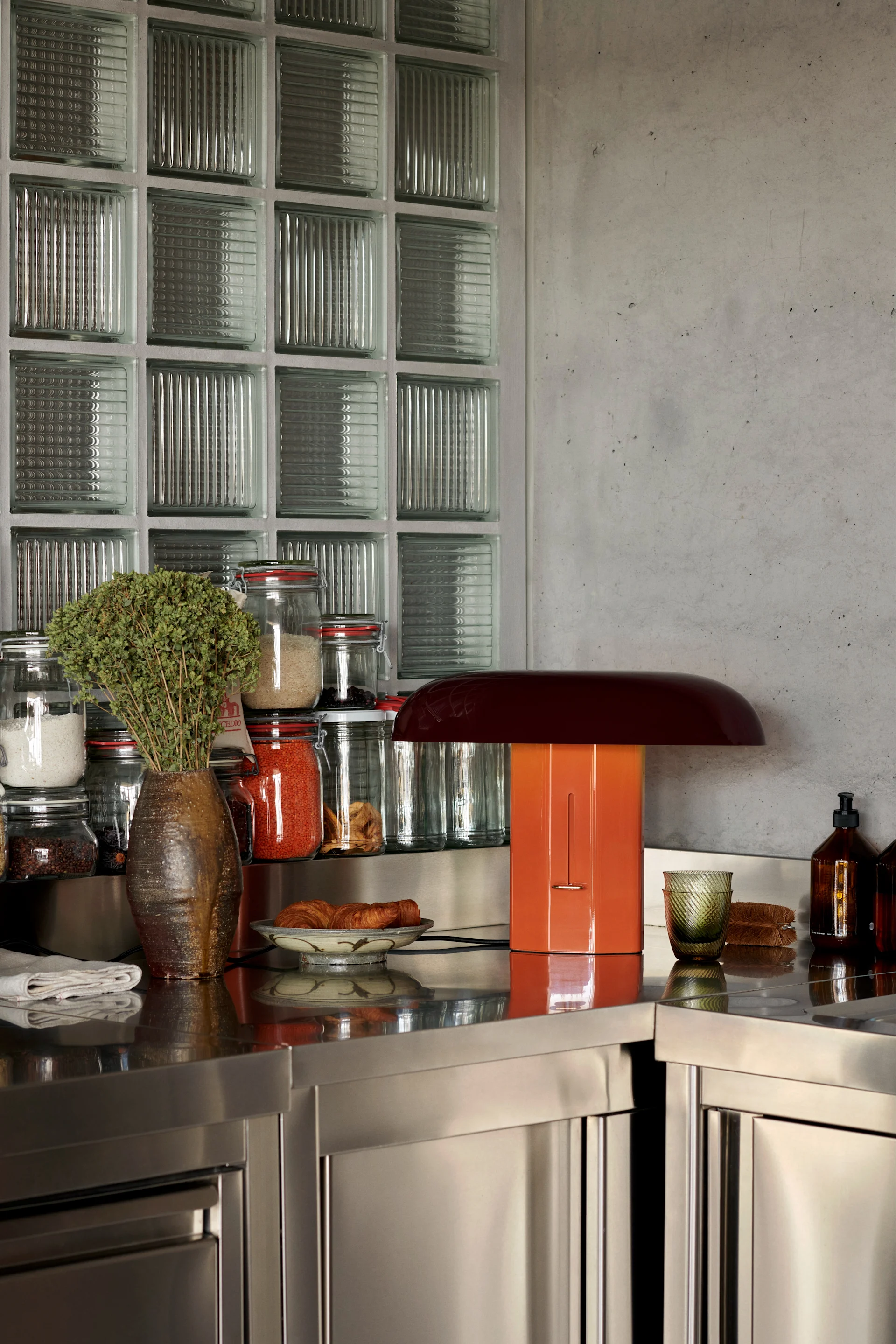 A stainless steel kitchen counter with a brown vase of herbs, jars of ingredients, an orange mushroom-shaped lamp, croissants, and glass bottles.