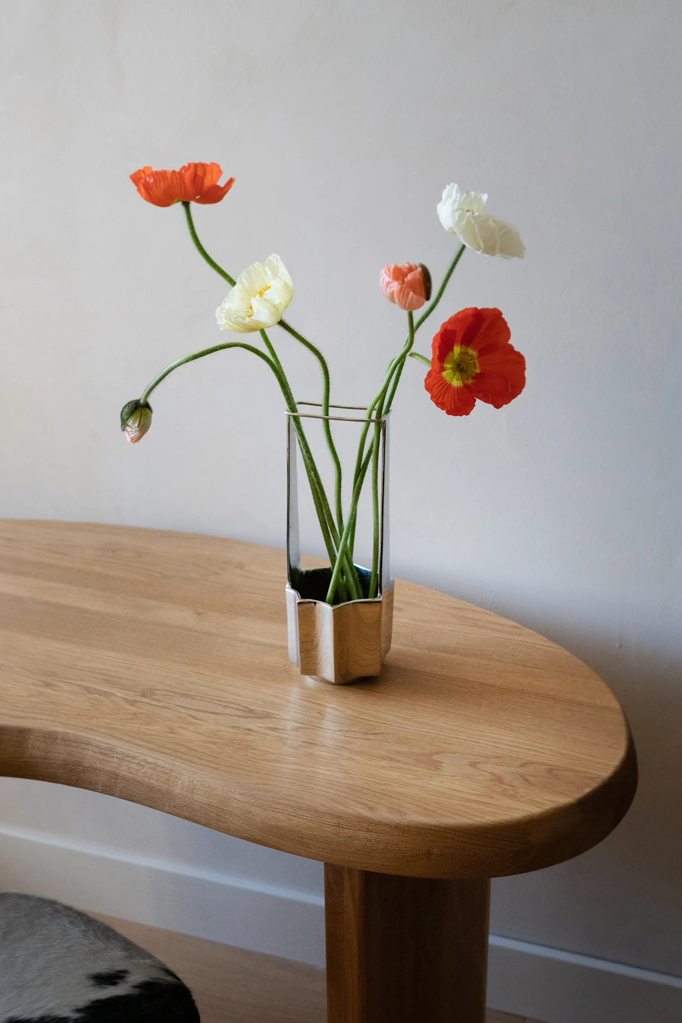 A bouquet of orange, yellow, and white poppies with long green stems in a unique glass and silver vase on a light wood table.