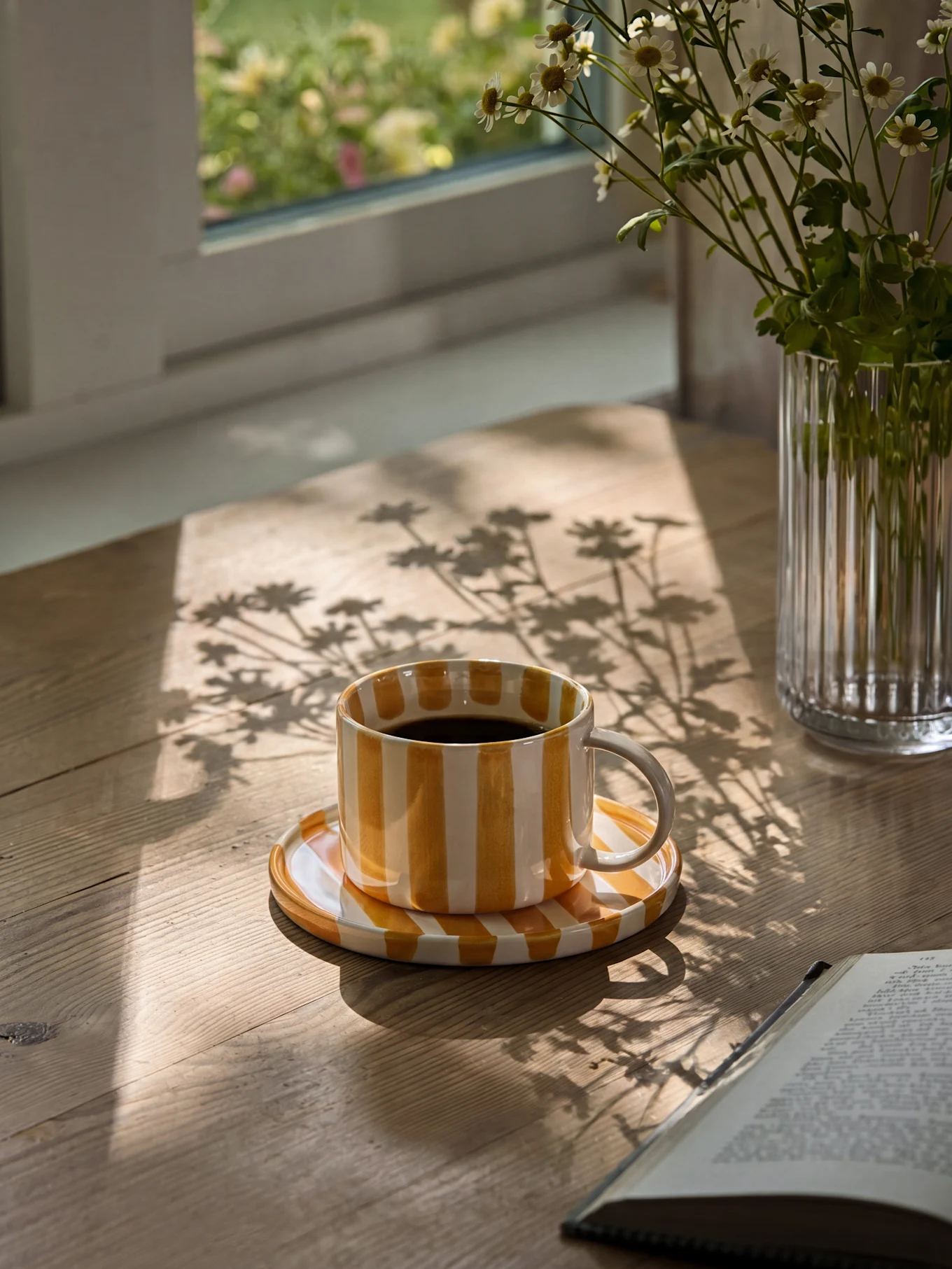 Yellow and white striped coffee cup with black coffee on a wooden table with sun shadows and a vase of daisies.
