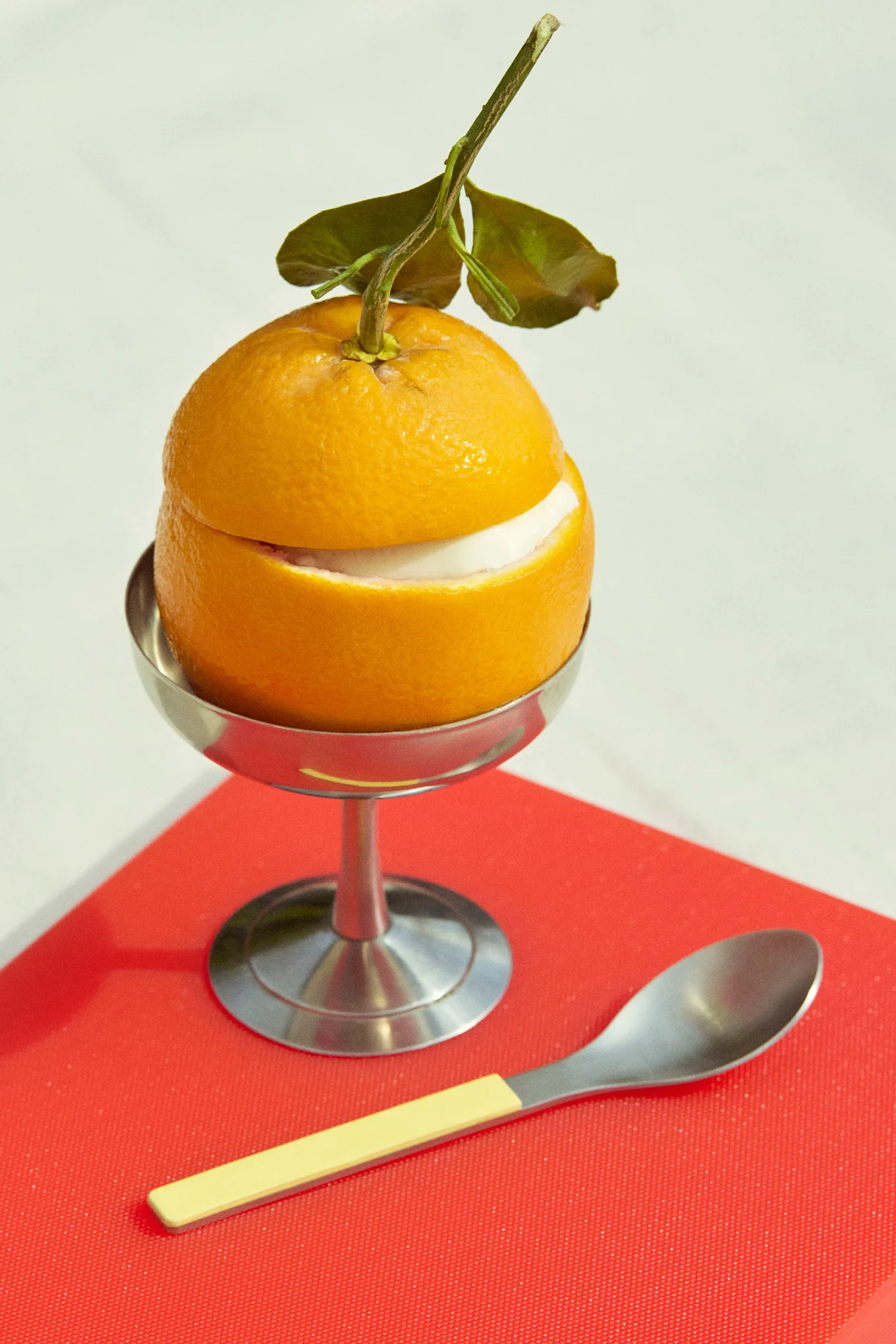 Orange dessert with white filling in a silver cup, with a spoon, on a red surface.