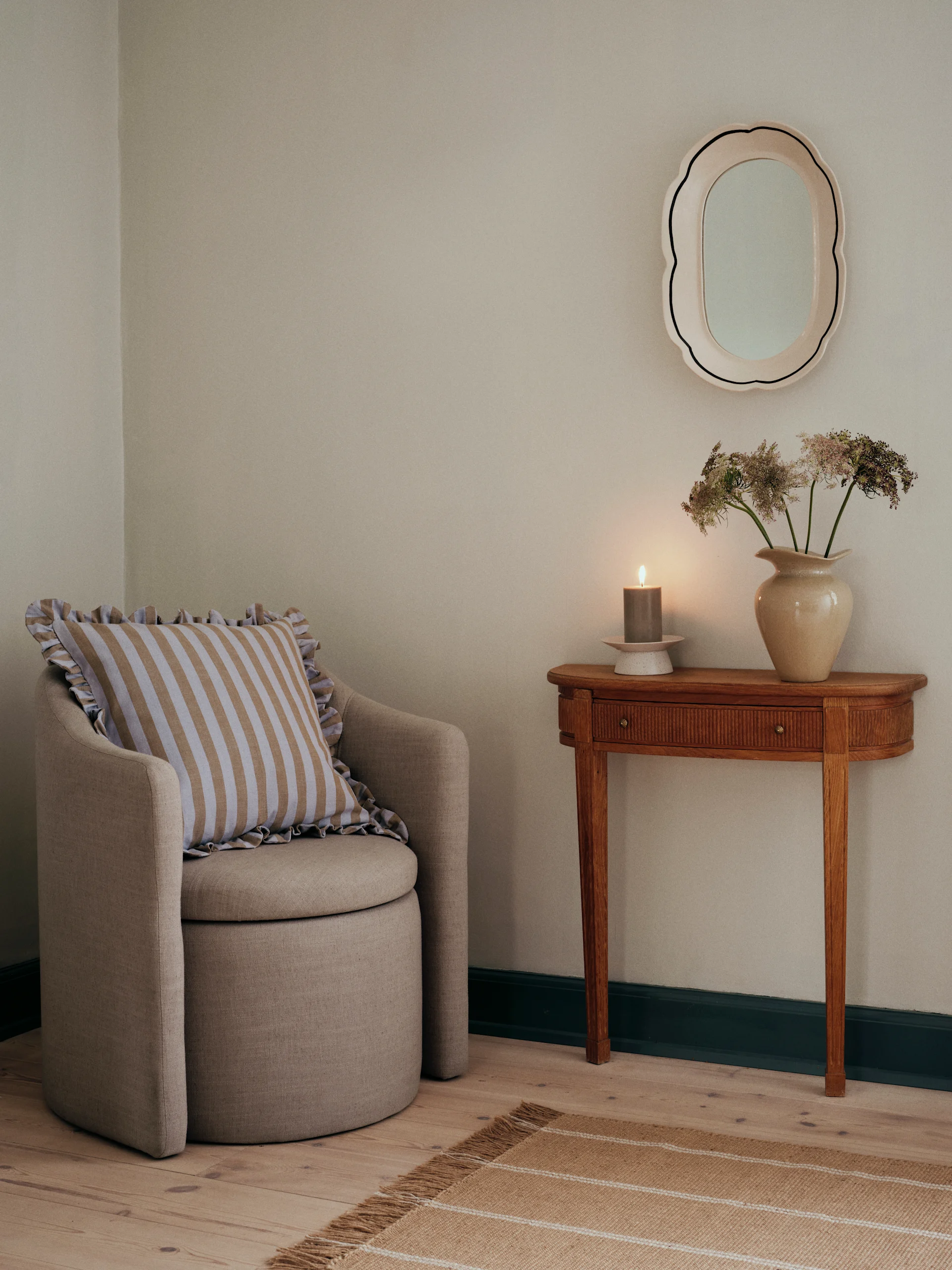 An armchair with a striped pillow next to a wooden console table with a lit candle, dried flowers, and a scalloped mirror.