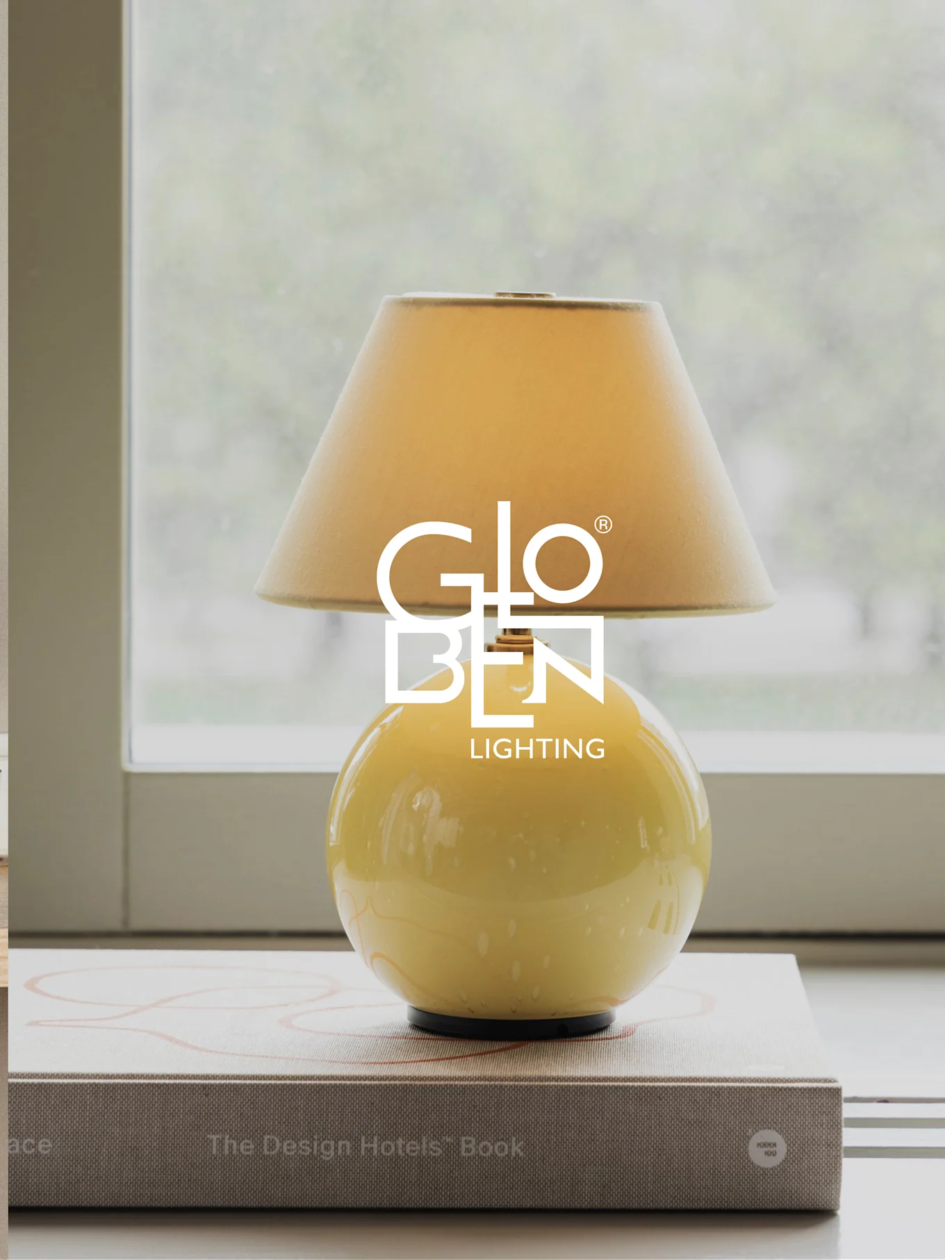 Yellow table lamp with a beige shade, sitting on books by a window.