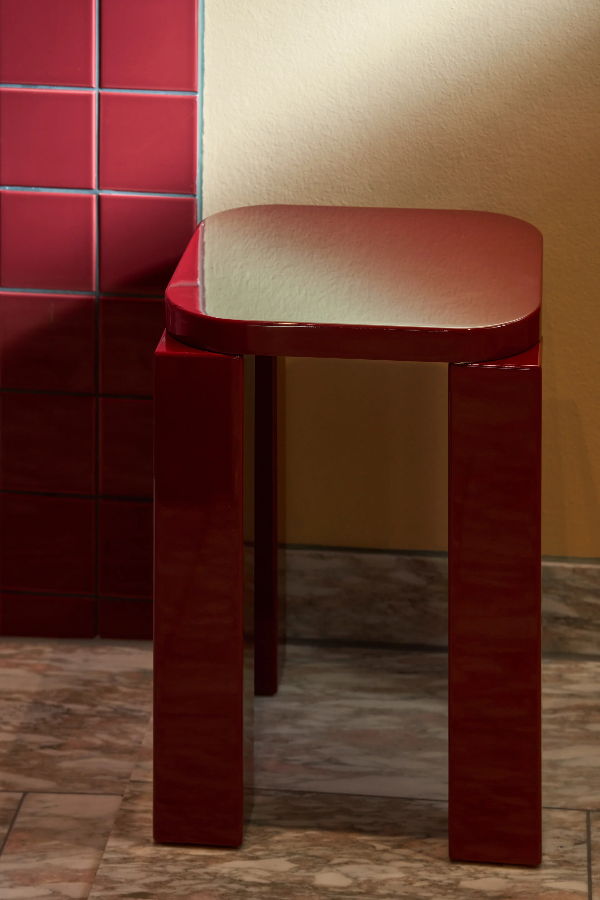 A shiny dark red stool with a rectangular top and four legs sits on a marble floor next to a tiled wall.