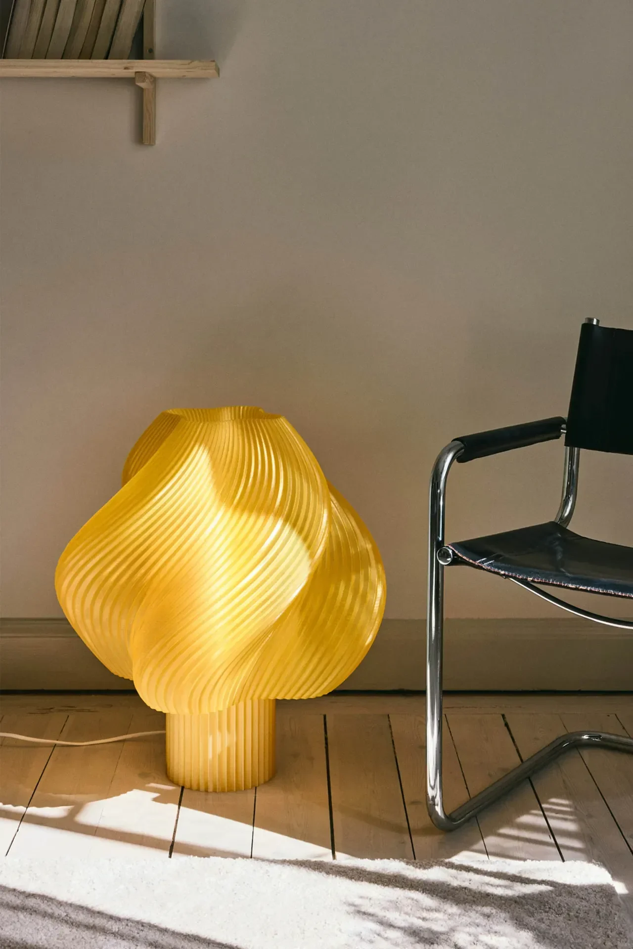 A bright yellow, sculptural table lamp with a ribbed, wavy design glows on a wooden floor next to a black leather and chrome chair.