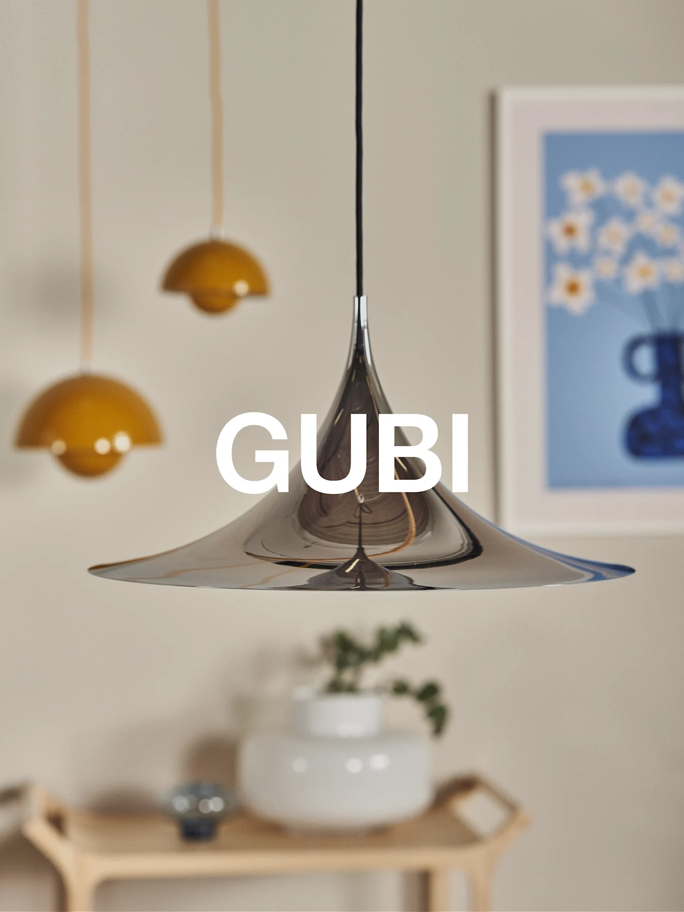 A polished chrome conical pendant lamp with the GUBI logo, set against a blurred background with other lamps and art.
