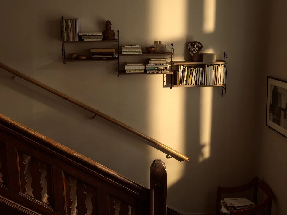 Sunlit wall with wooden shelves holding books and decor, alongside a wooden staircase and handrail.