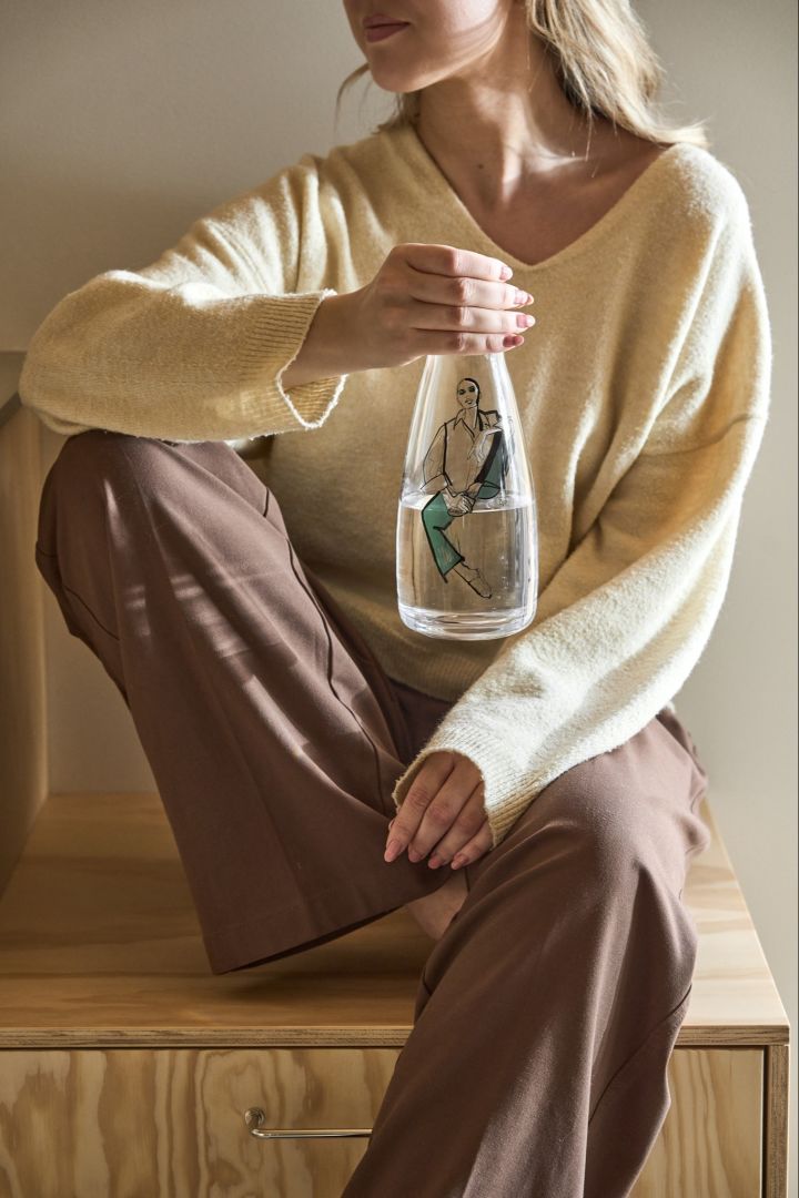 A woman sits with her knee up and holding the Time for you Carafe from the All about you collection from Kosta Boda.